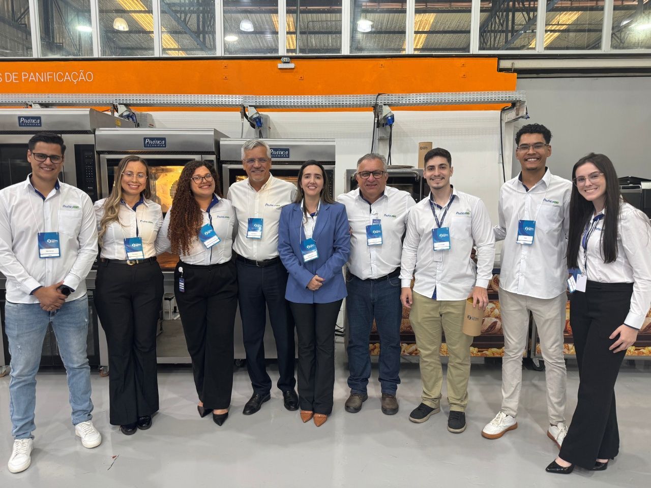Grupo de pessoas sorrindo em uma fábrica. Algumas usam cordões. Máquinas e placas laranja ao fundo.