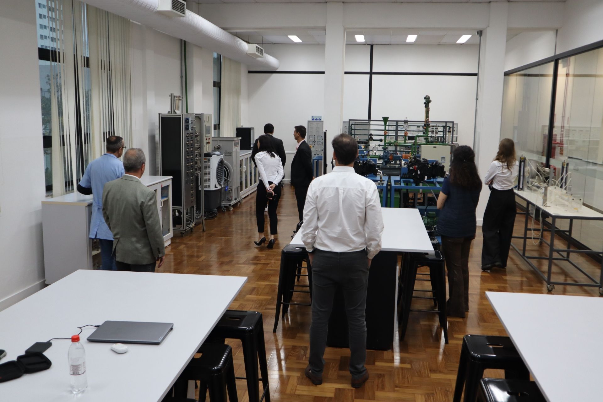 Pessoas visitando um laboratório industrial com máquinas, mesas brancas e janelas.