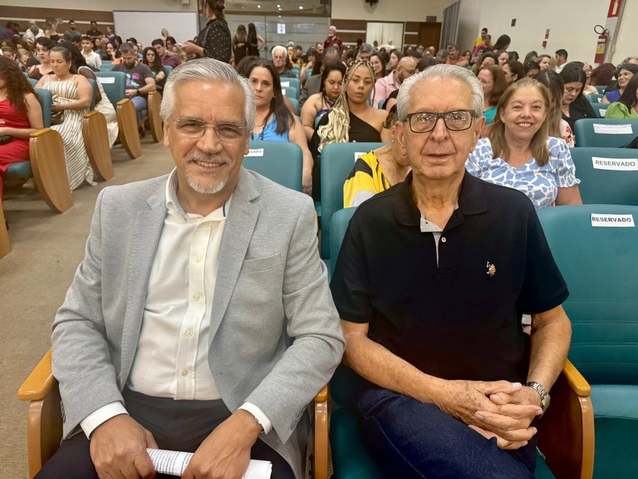 Dois homens estão sentados em cadeiras em frente a uma multidão de pessoas.
