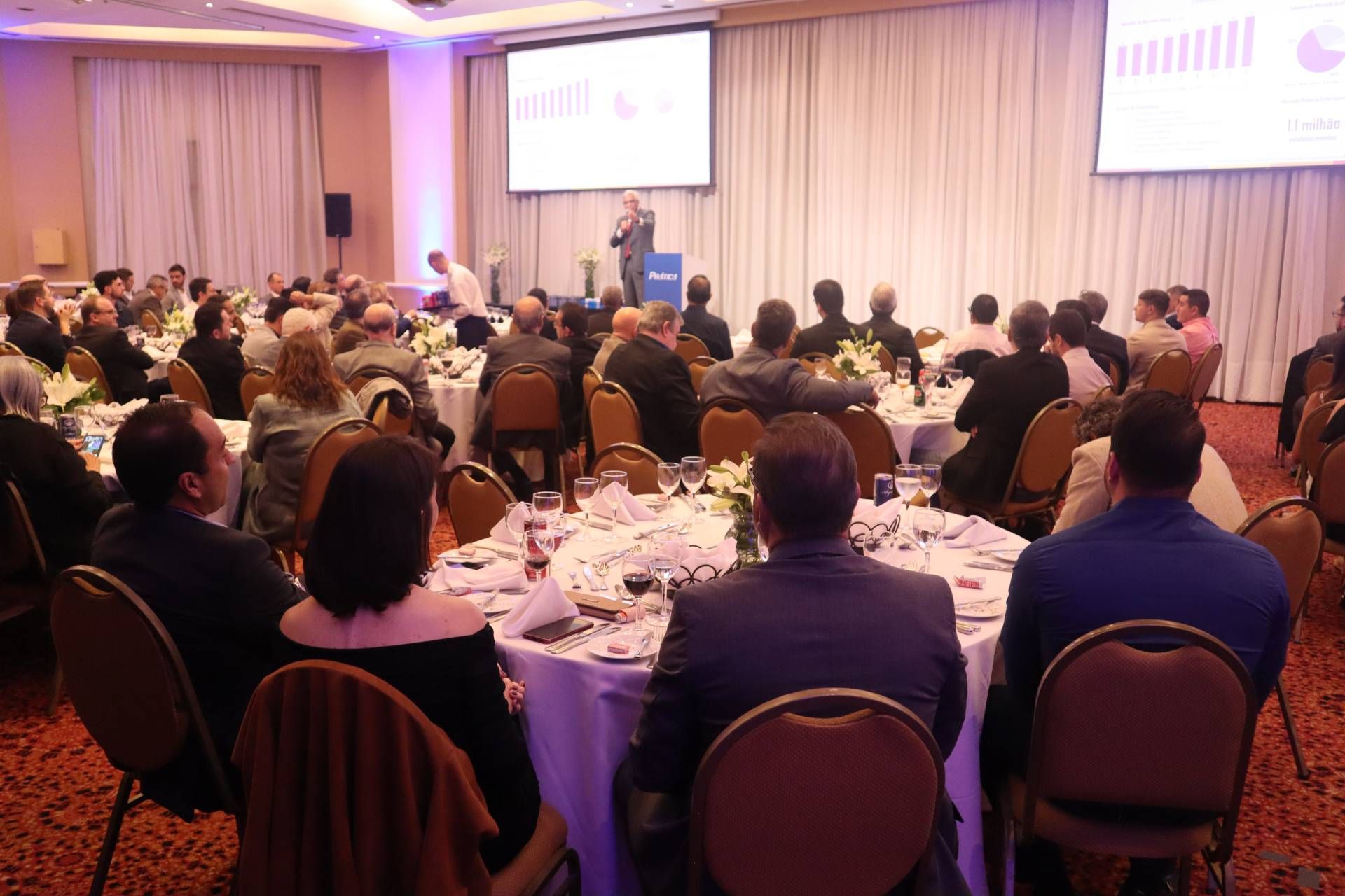 Um grupo de pessoas está sentado em mesas em uma sala grande assistindo a uma apresentação.