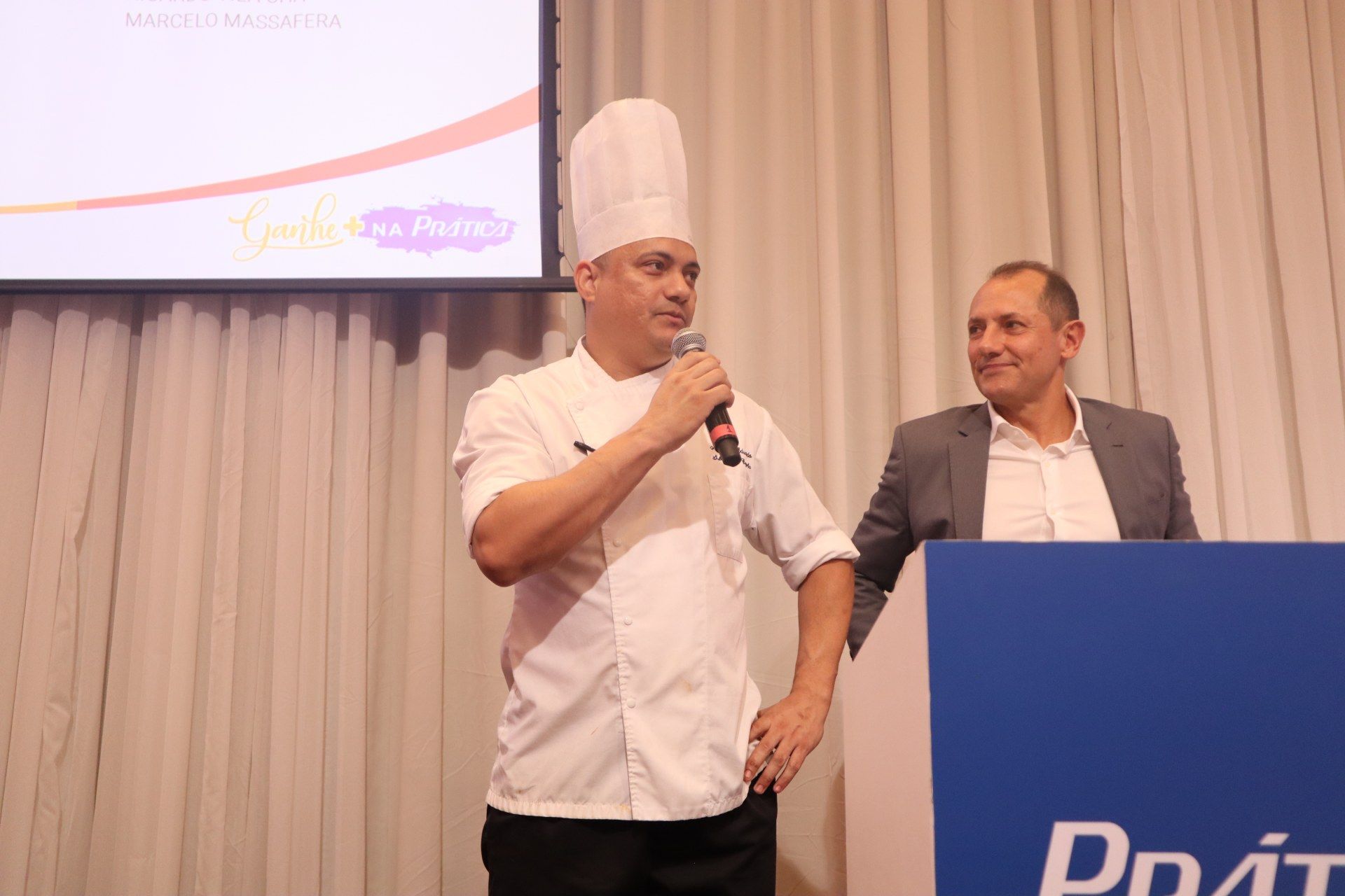 Um chef está falando em um microfone enquanto está ao lado de um homem de terno.