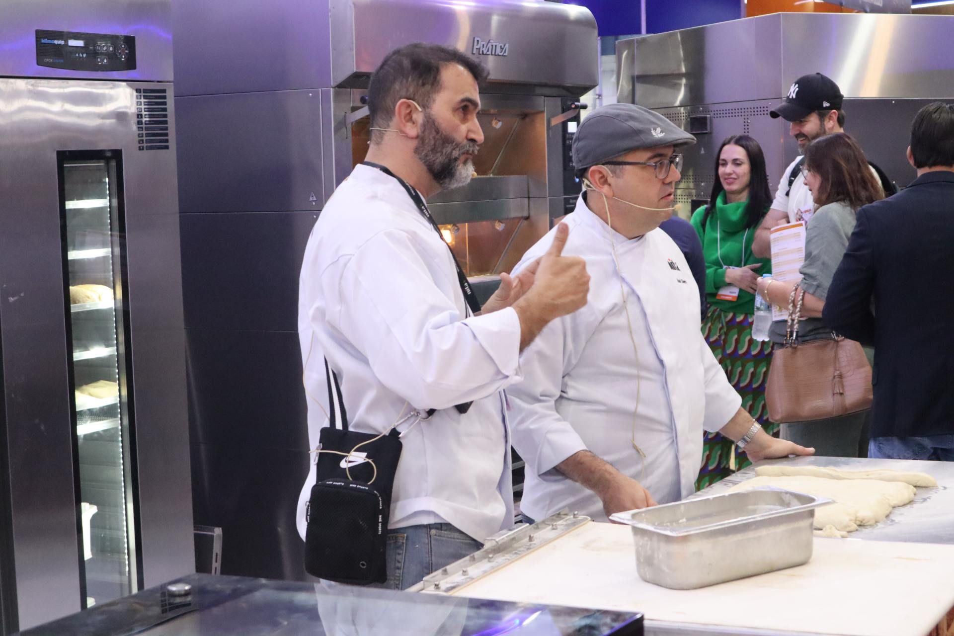 Dois homens estão de pé, um ao lado do outro, em uma cozinha, e um deles está fazendo sinal de positivo.