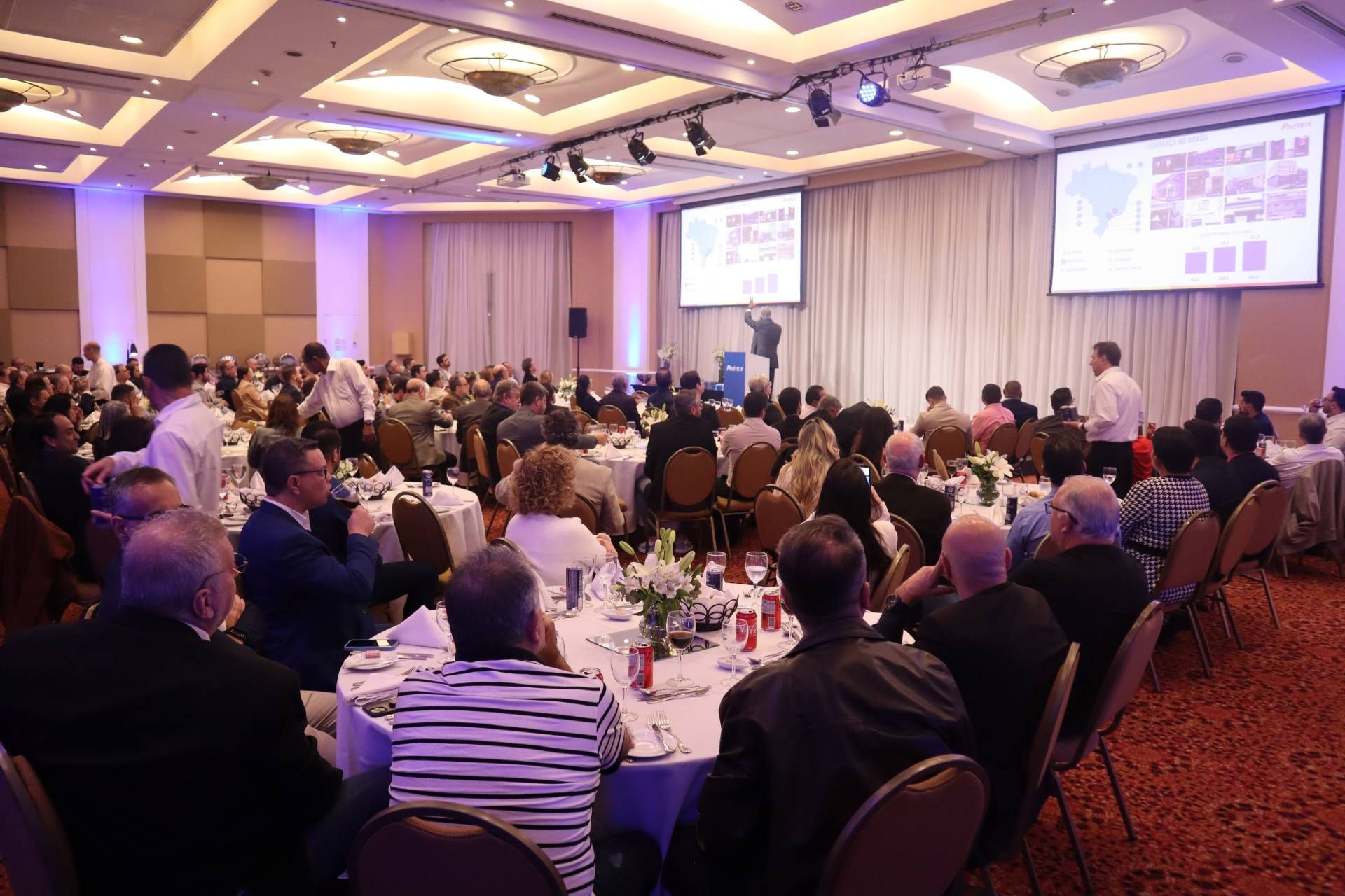 Um grande grupo de pessoas está sentado em mesas em uma sala grande.