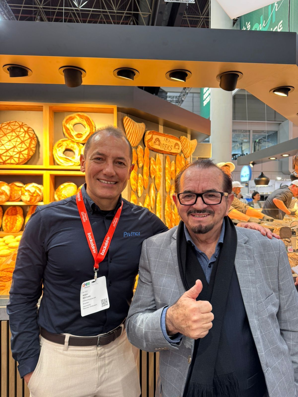 Dois homens parados um ao lado do outro em frente a uma padaria fazendo sinal de positivo