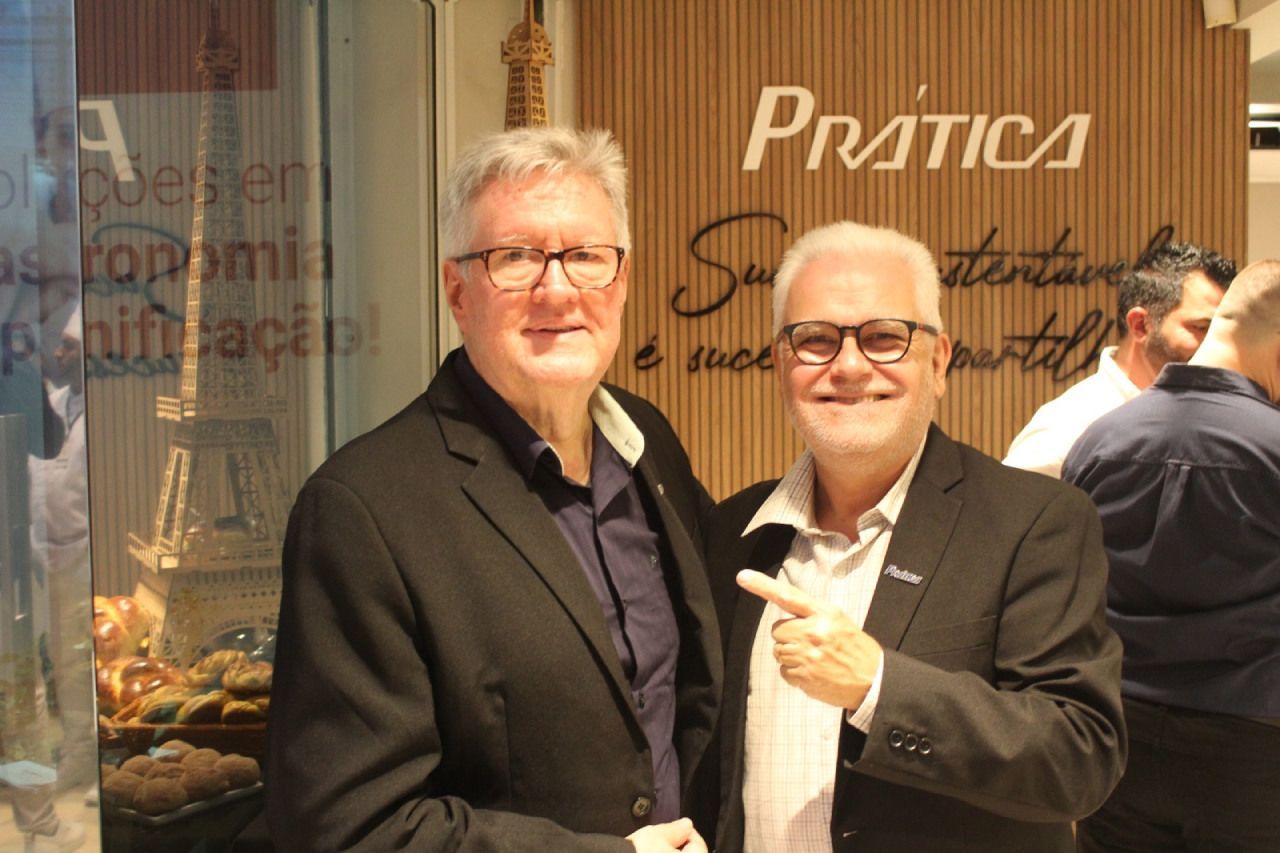 Dois homens posam para uma foto em frente a uma placa que diz pratica