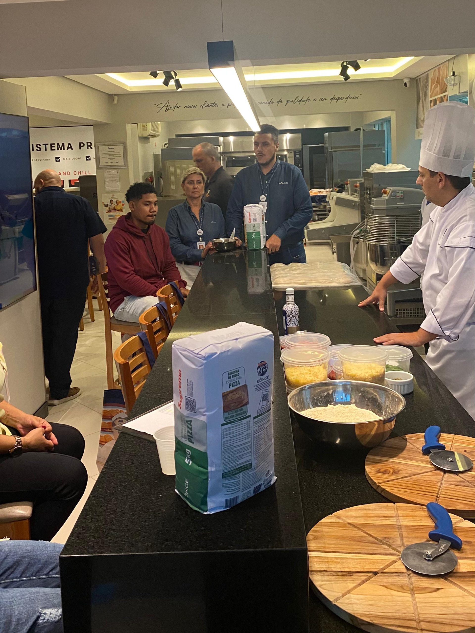 Um grupo de pessoas está sentado à mesa em uma cozinha enquanto um chef prepara comida.