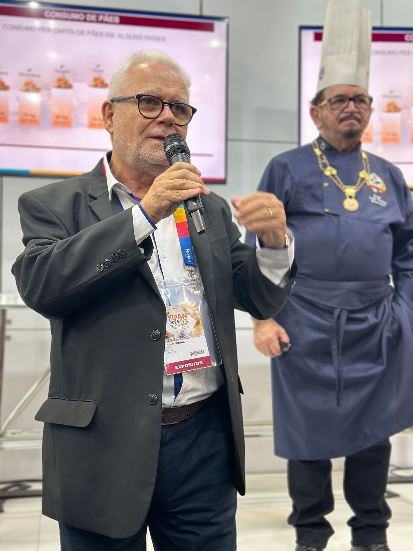 Um homem de terno fala em um microfone enquanto um chef está atrás dele.