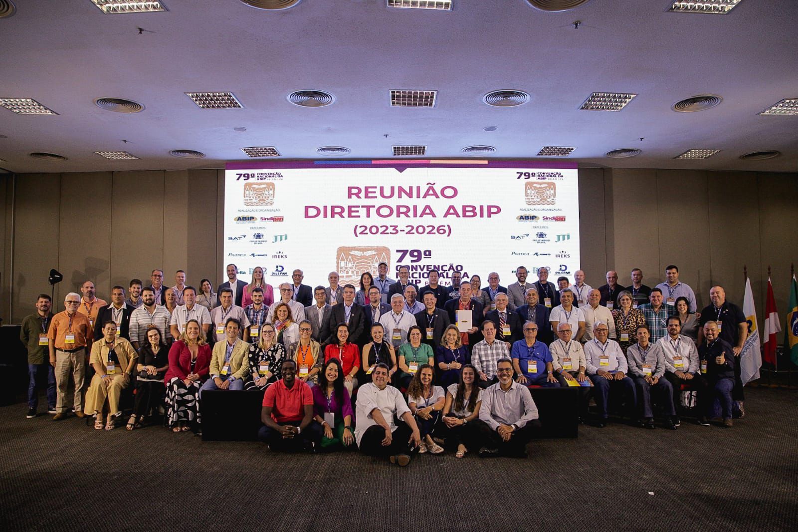 Um grande grupo de pessoas está posando para uma foto em frente a uma tela grande.