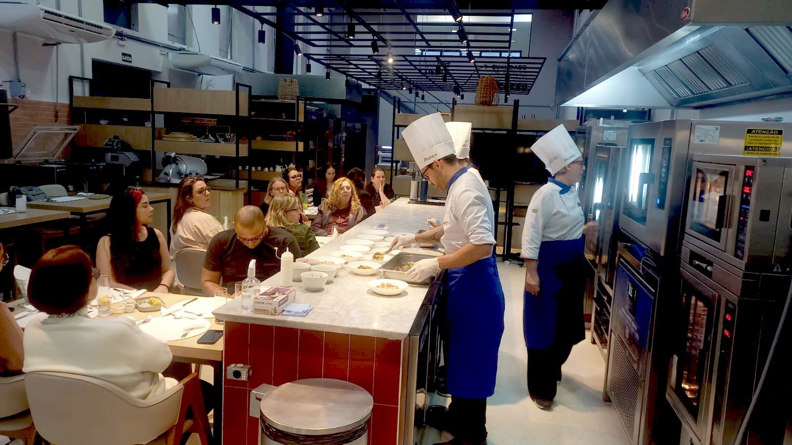 Chef montando pratos no balcão enquanto os clientes observam; ambiente de cozinha industrial.