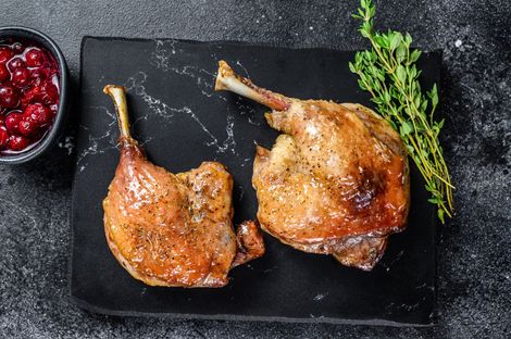 Duas coxas de pato assadas e douradas com ervas, servidas sobre uma laje de pedra preta, ao lado de uma tigela de molho de frutos vermelhos.