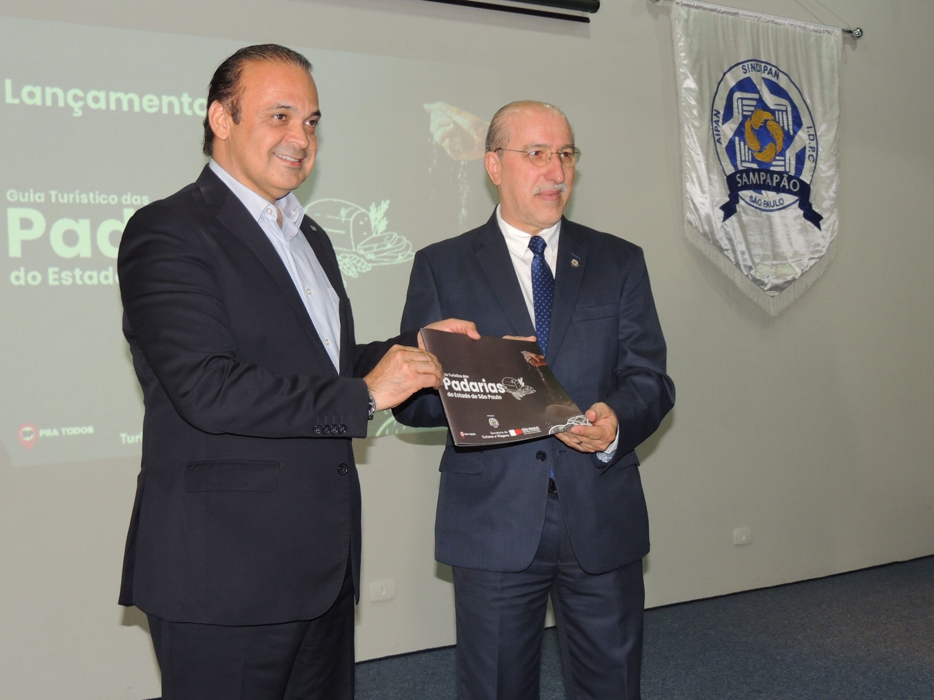 Dois homens de terno estão parados um ao lado do outro e um deles está segurando um livro