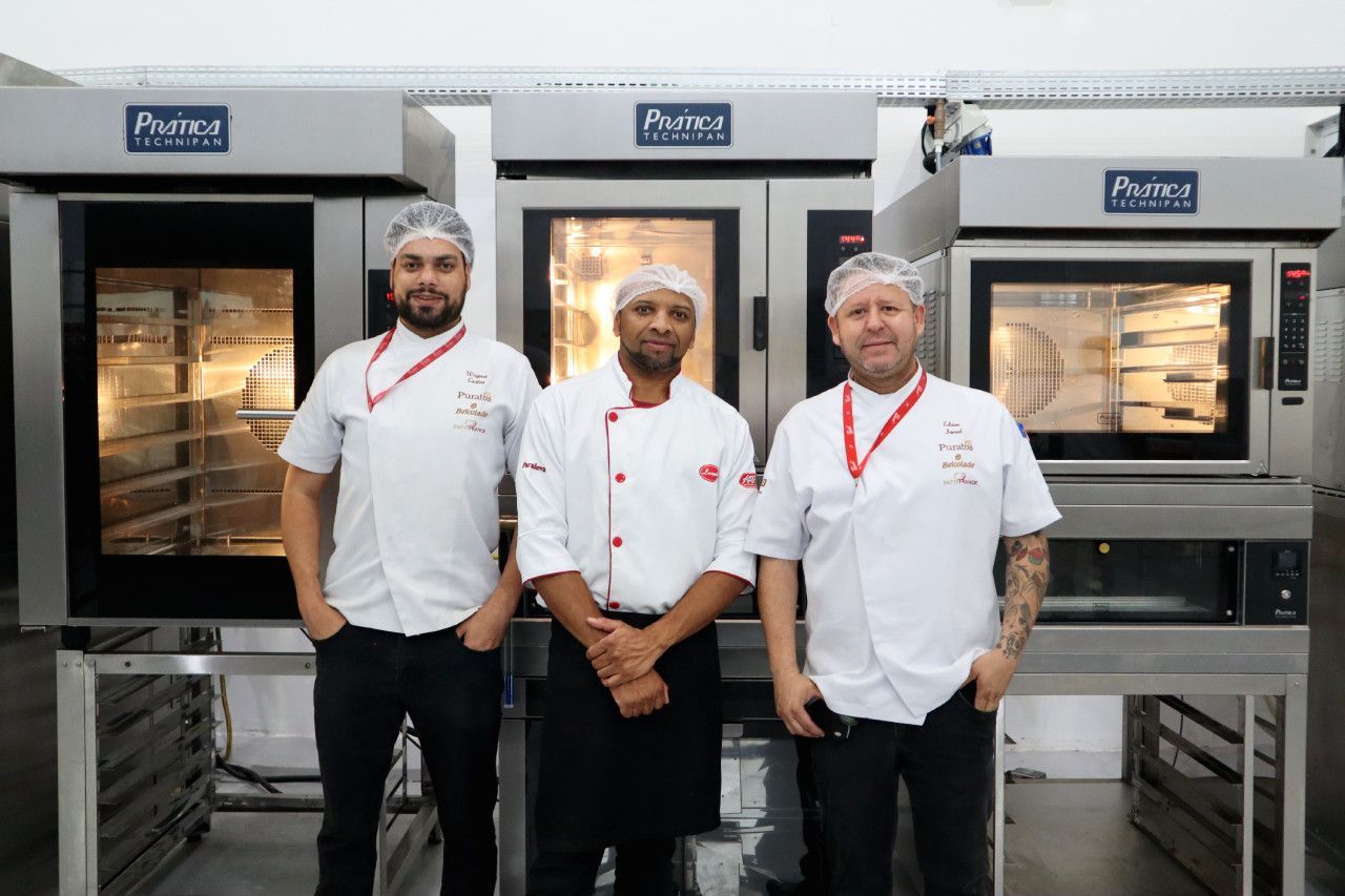 Três homens estão de pé, um ao lado do outro, em frente aos fornos de uma cozinha.