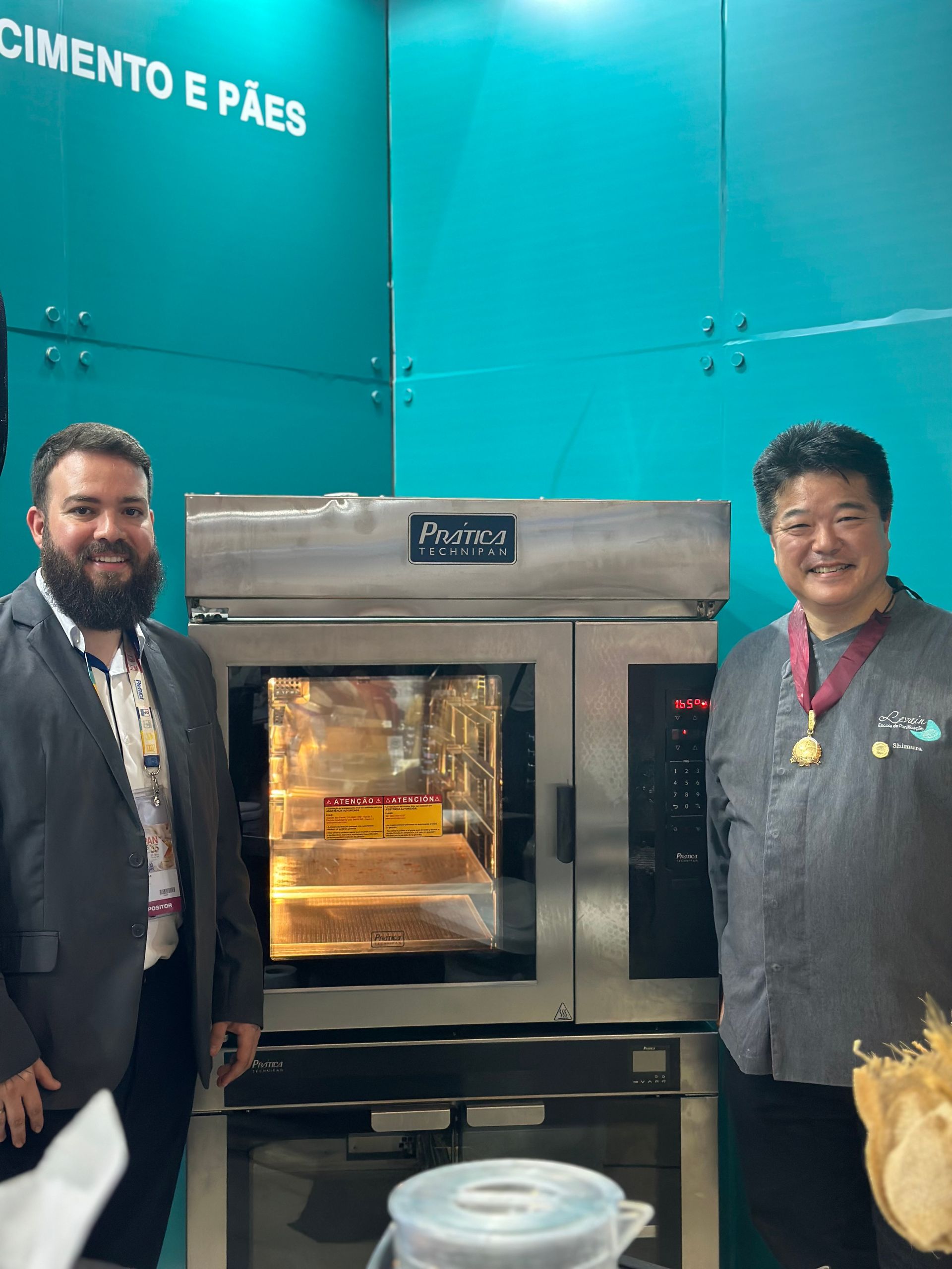 Dois homens em pé em frente a um forno com a palavra cimento e pão na parede atrás deles