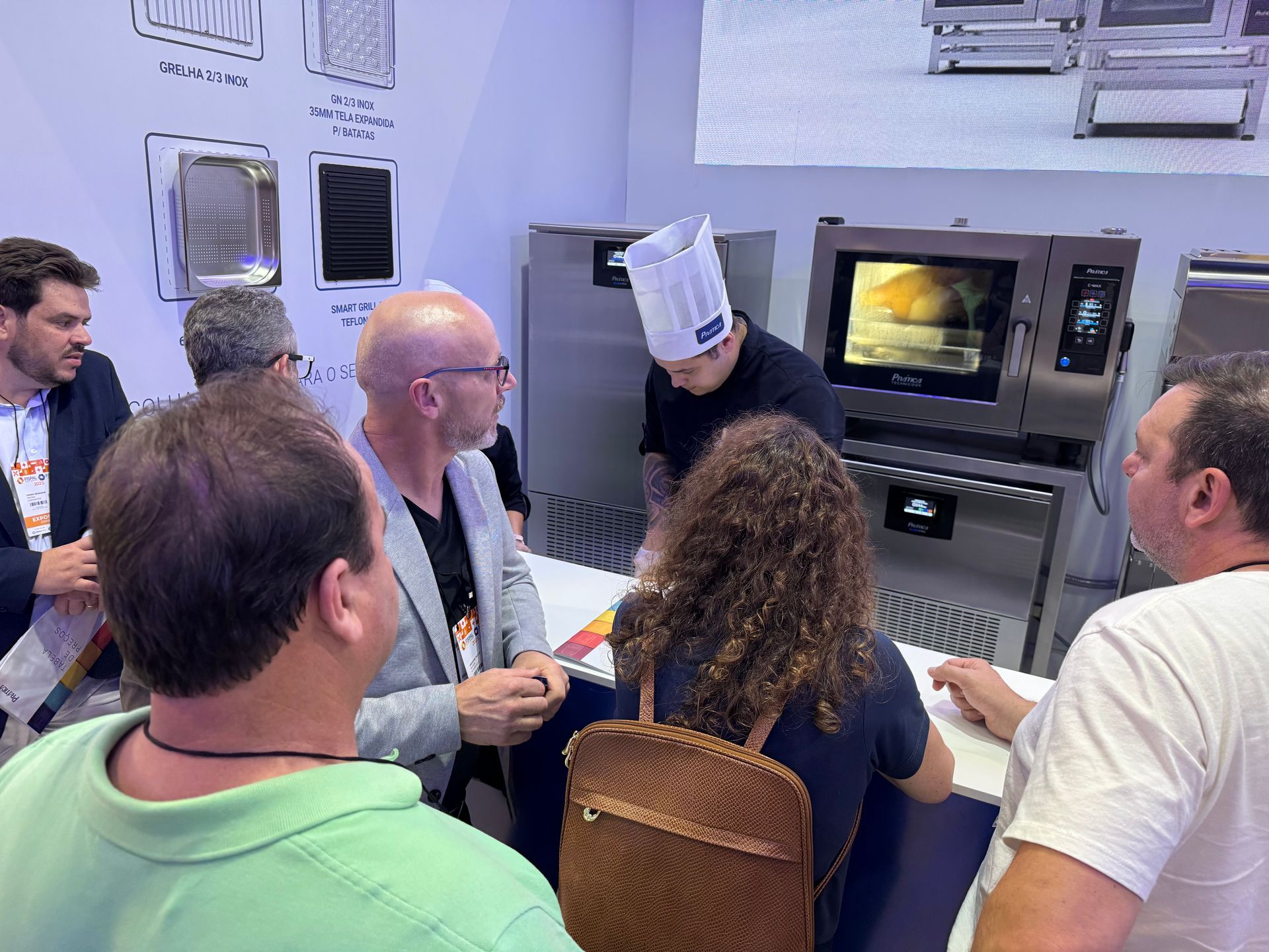 Um grupo de pessoas está sentado ao redor de uma mesa conversando com um homem com chapéu de chef.