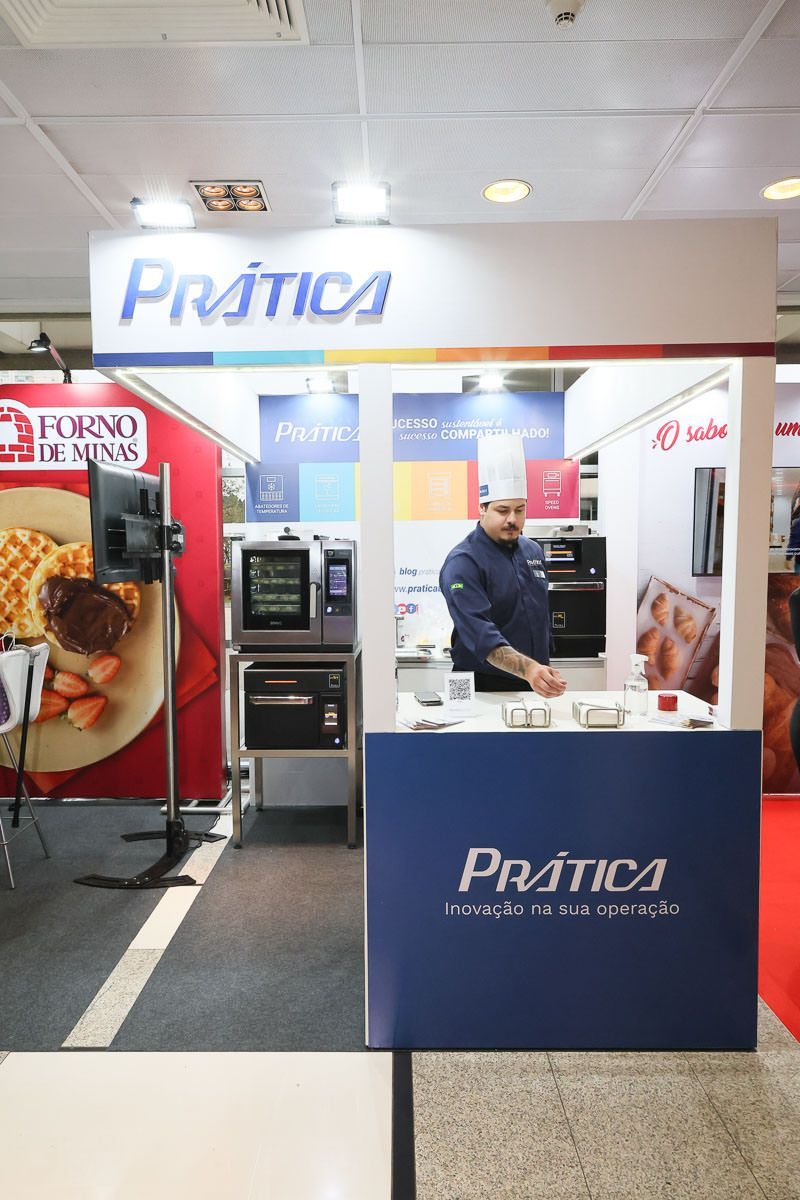 Chef no estande da Prática exibindo fornos. Marca azul e branca, ambiente de convenção.