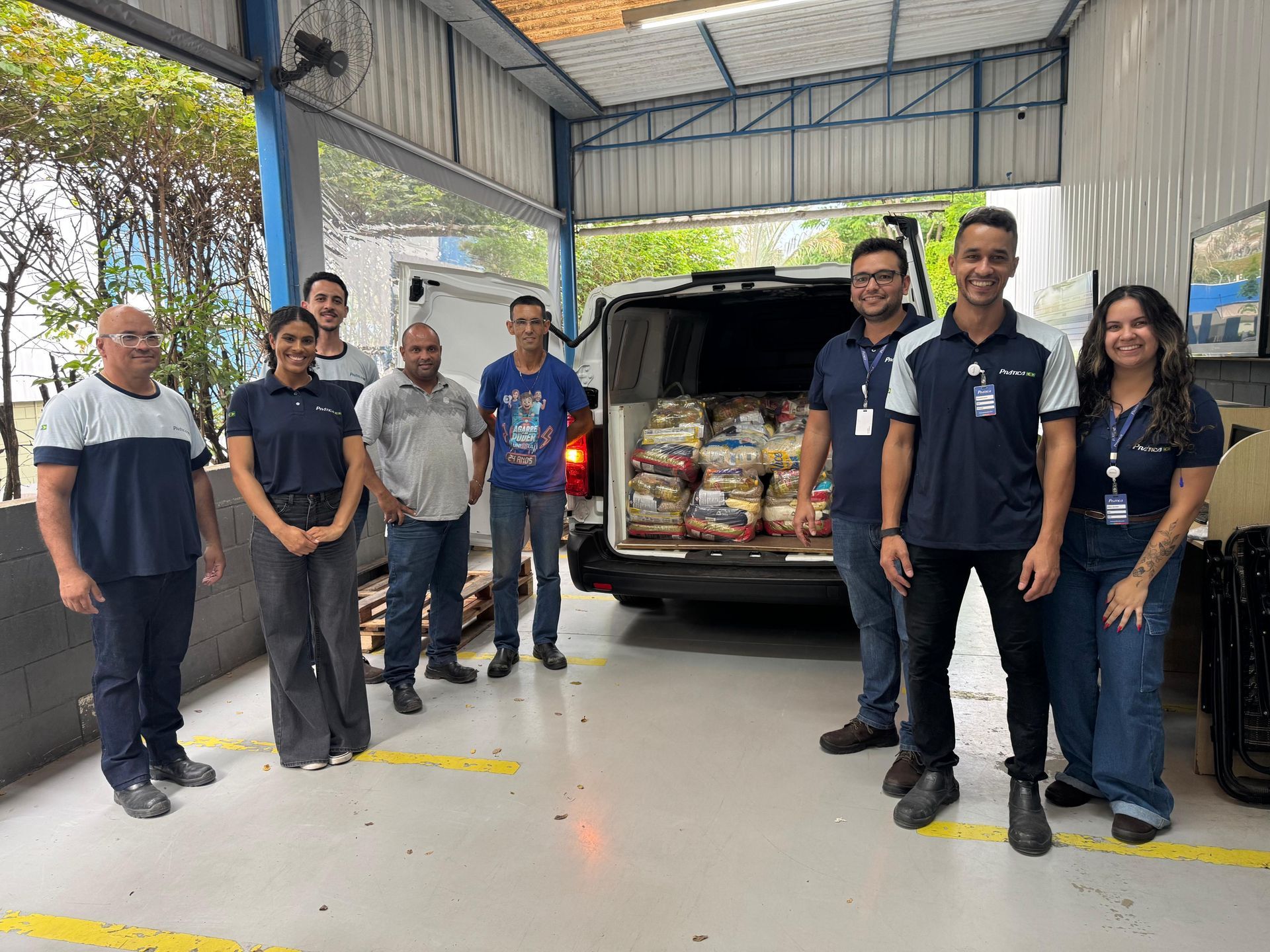 Grupo de pessoas carregando comida em uma van dentro de um armazém. Elas sorriem, e algumas usam camisetas combinando.