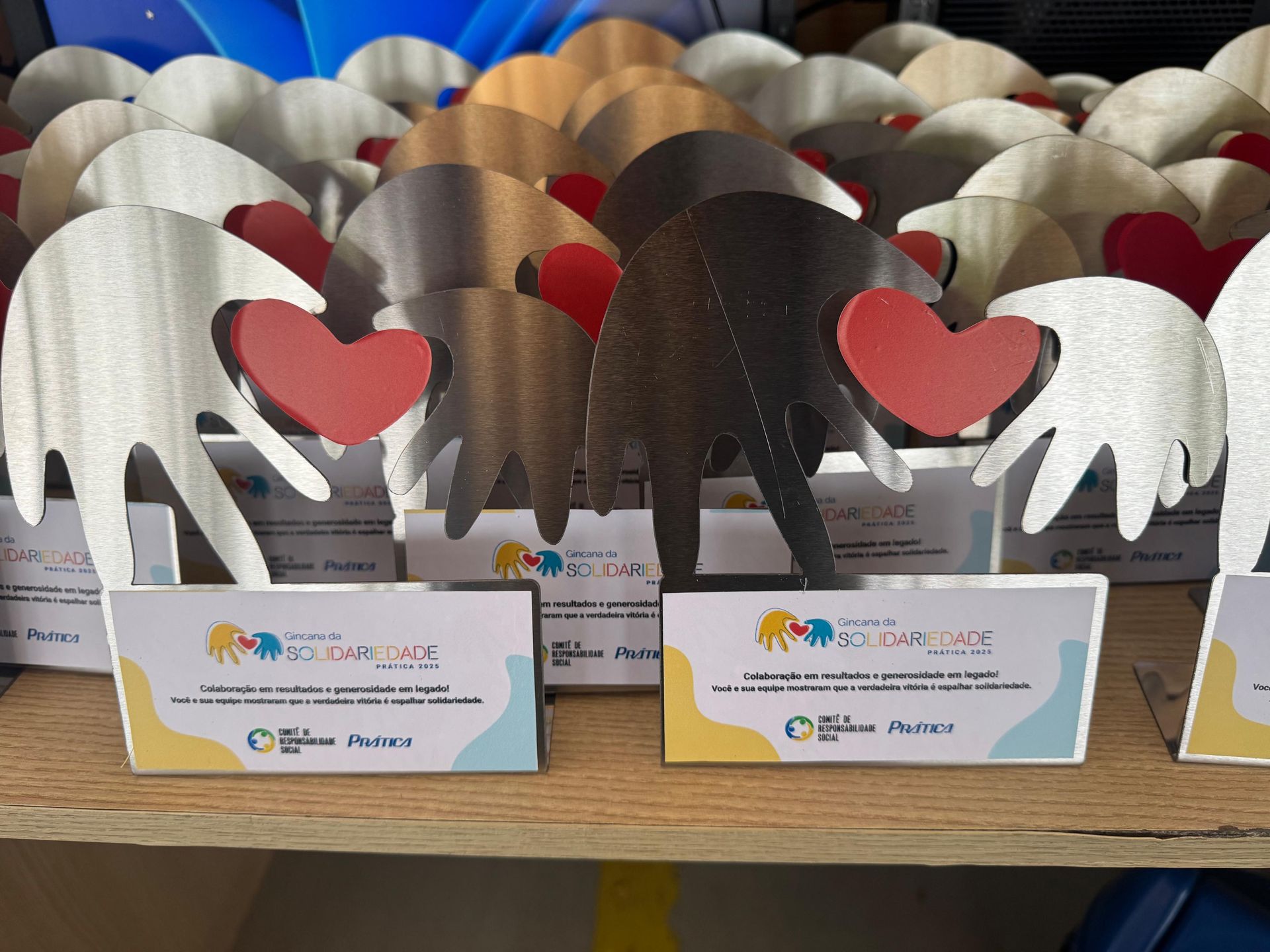 Prêmios com recortes de metal em formato de mãos segurando corações vermelhos; cada um tem uma placa de identificação com logotipo e texto.