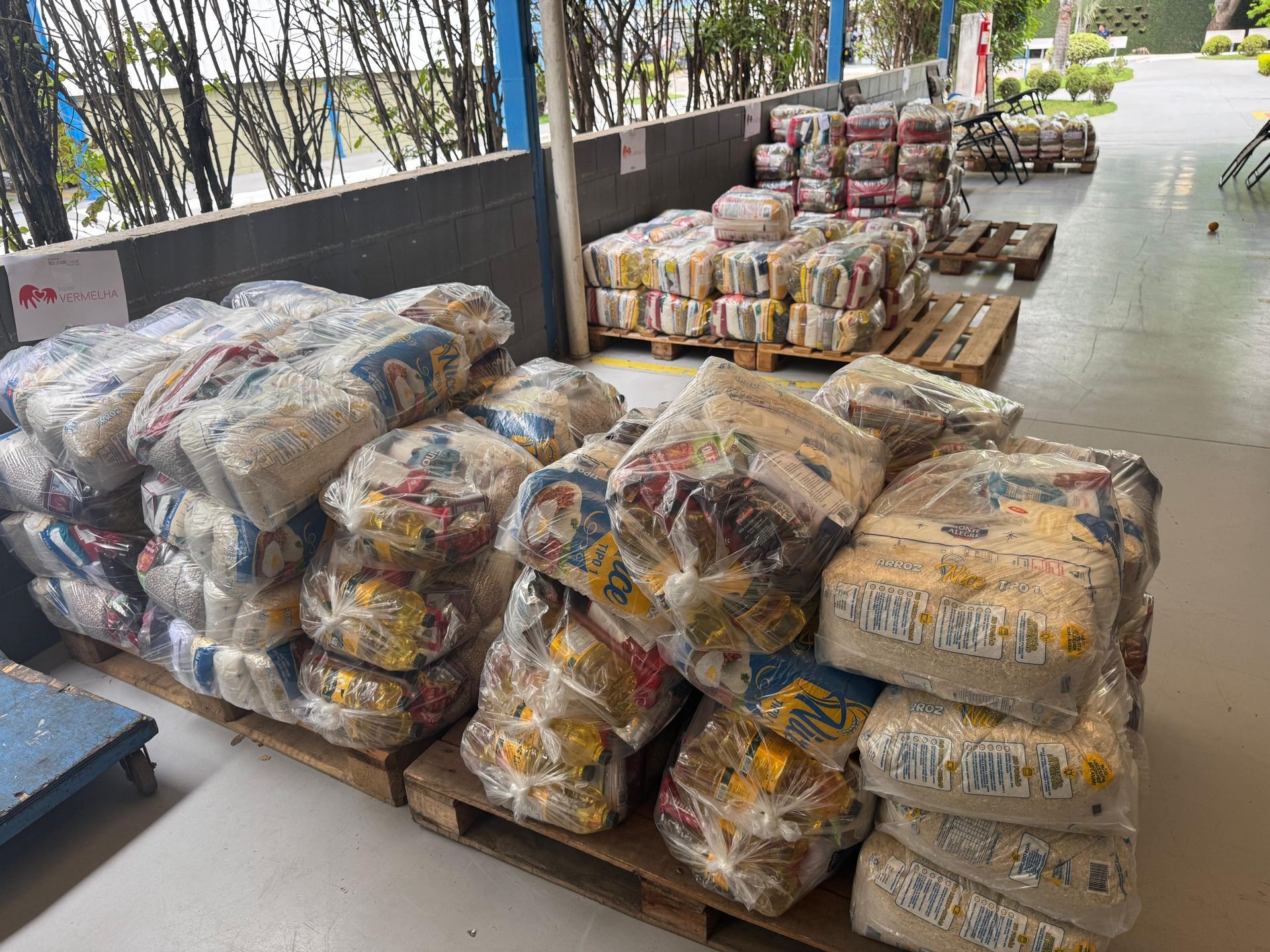 Sacos de comida, embrulhados em plástico, empilhados em paletes. Localizados dentro de casa. Outros, em paletes, atrás.
