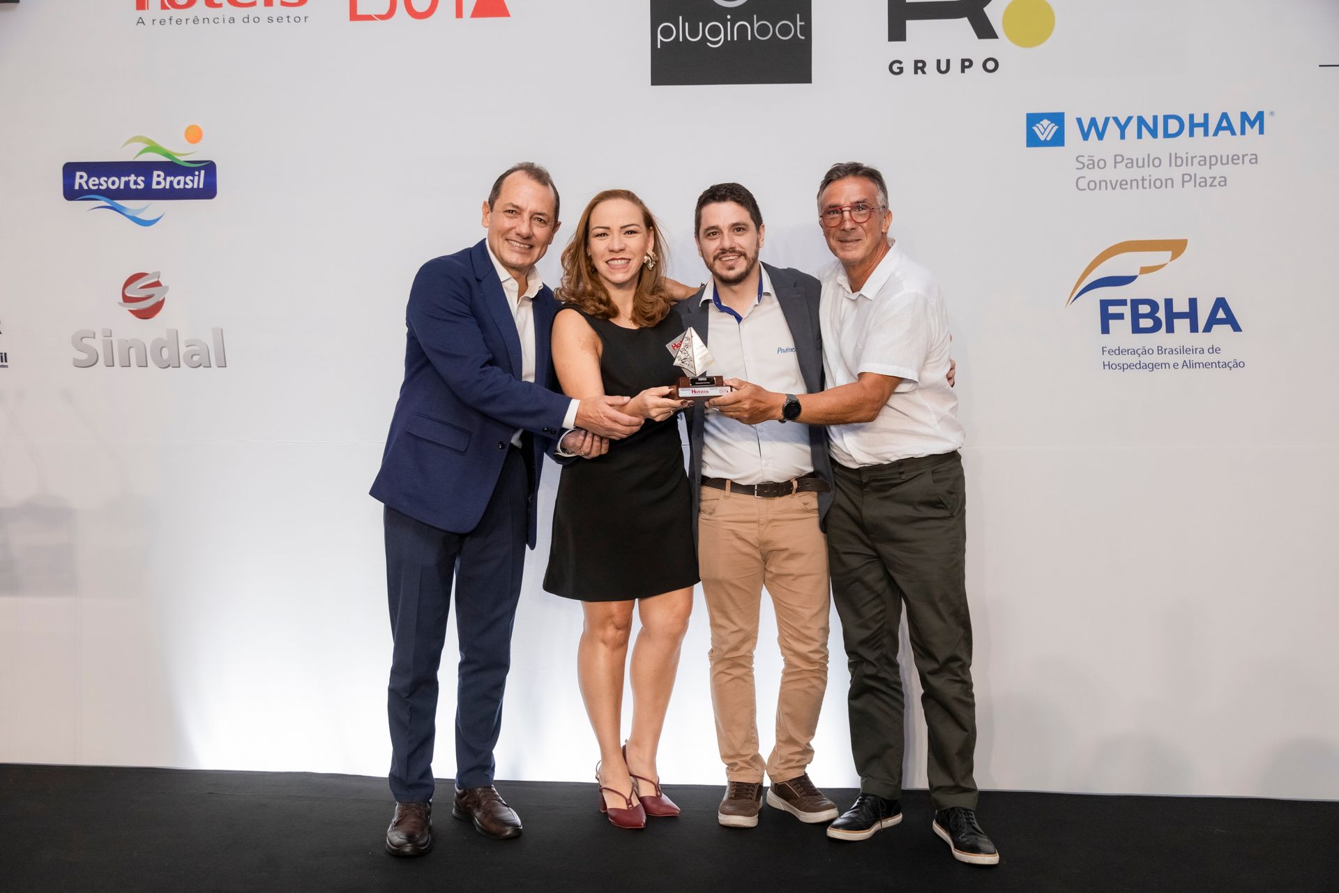 Um grupo de pessoas em pé, uma ao lado da outra, em frente a uma parede com logotipos.