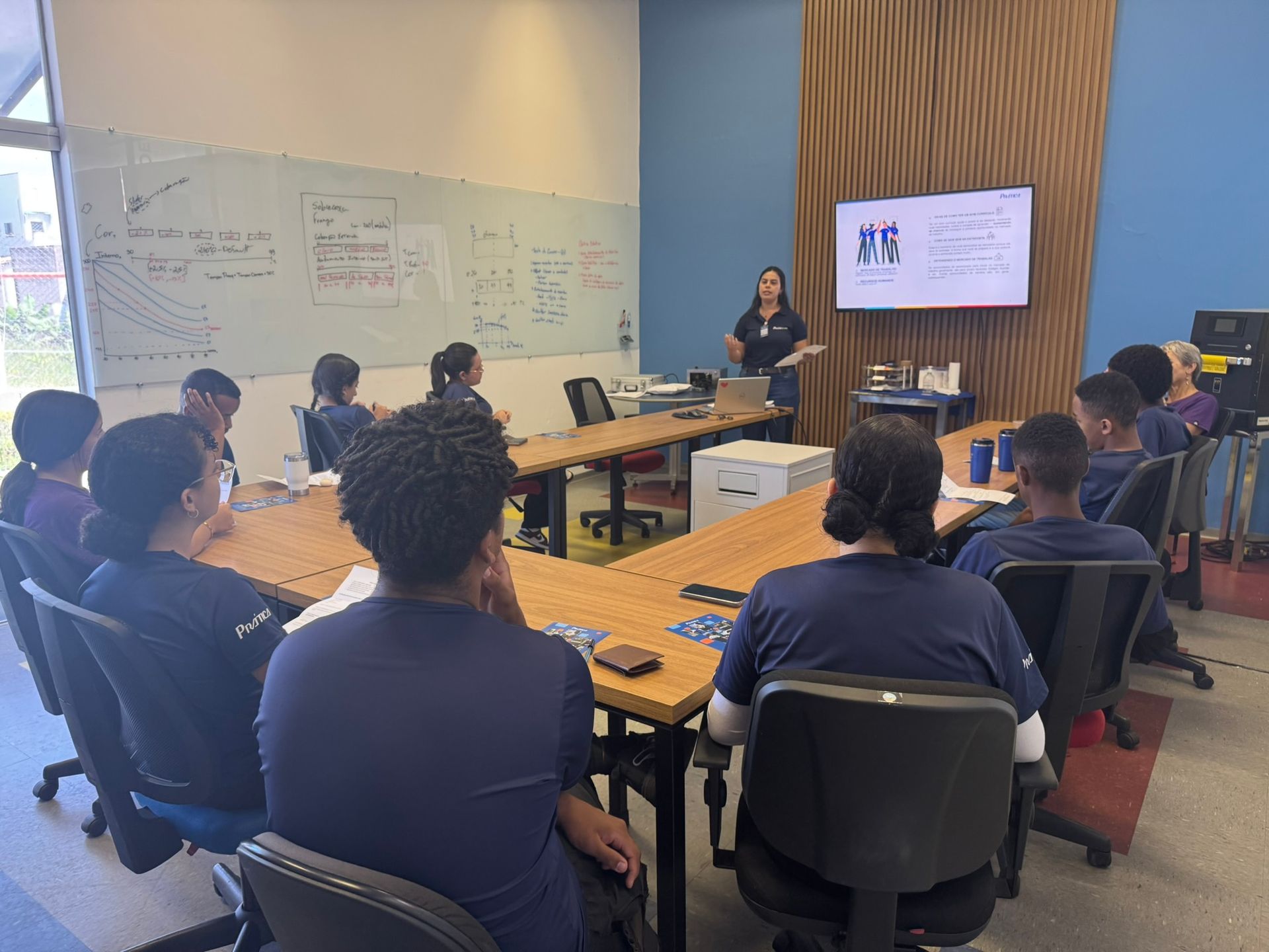 Uma mulher faz uma apresentação para um grupo de pessoas em uma sala de reuniões, com uma tela atrás dela.