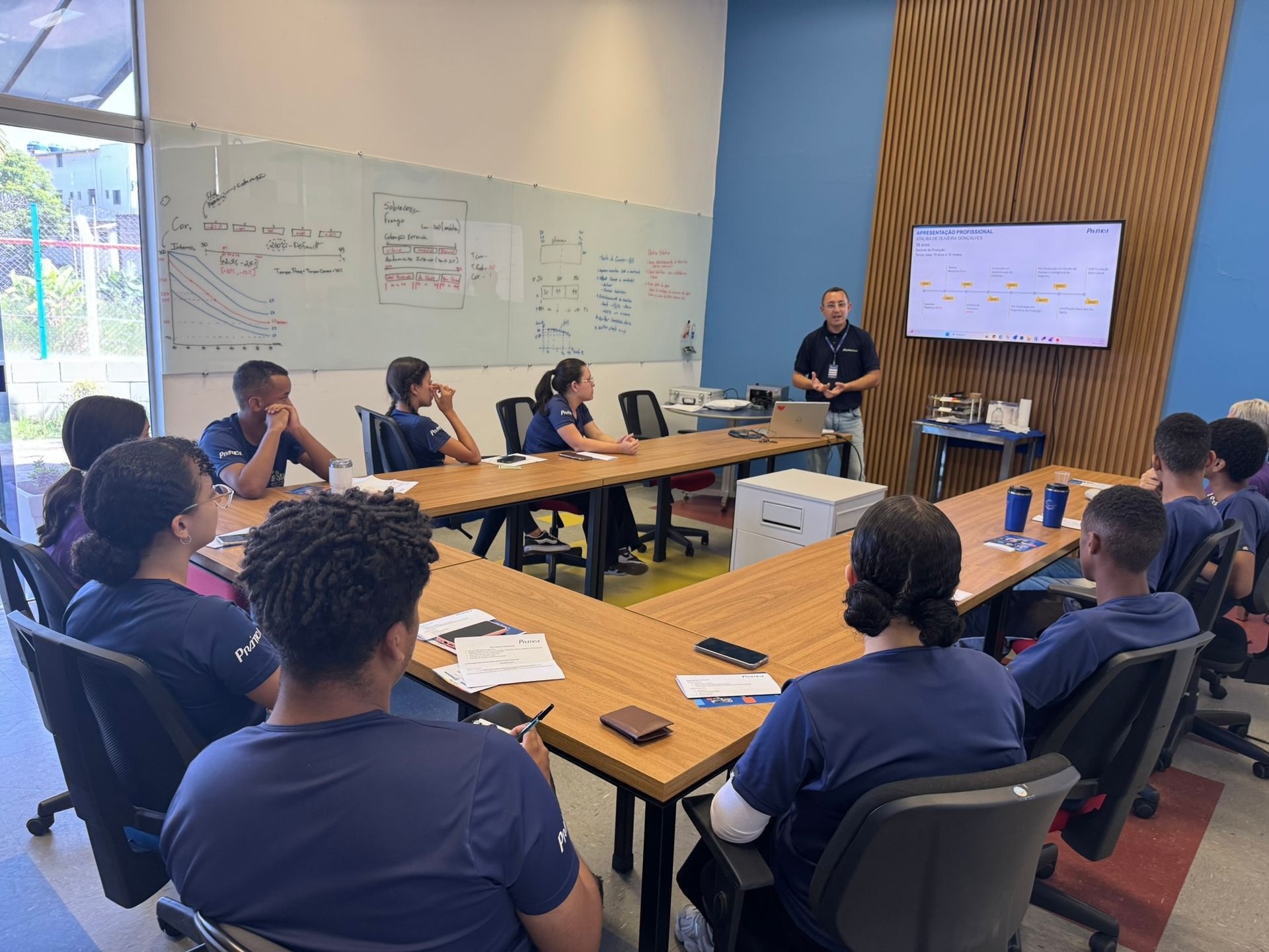 Um grupo de pessoas vestindo camisas azuis está reunido. Um homem está na frente, apresentando algo em uma tela.