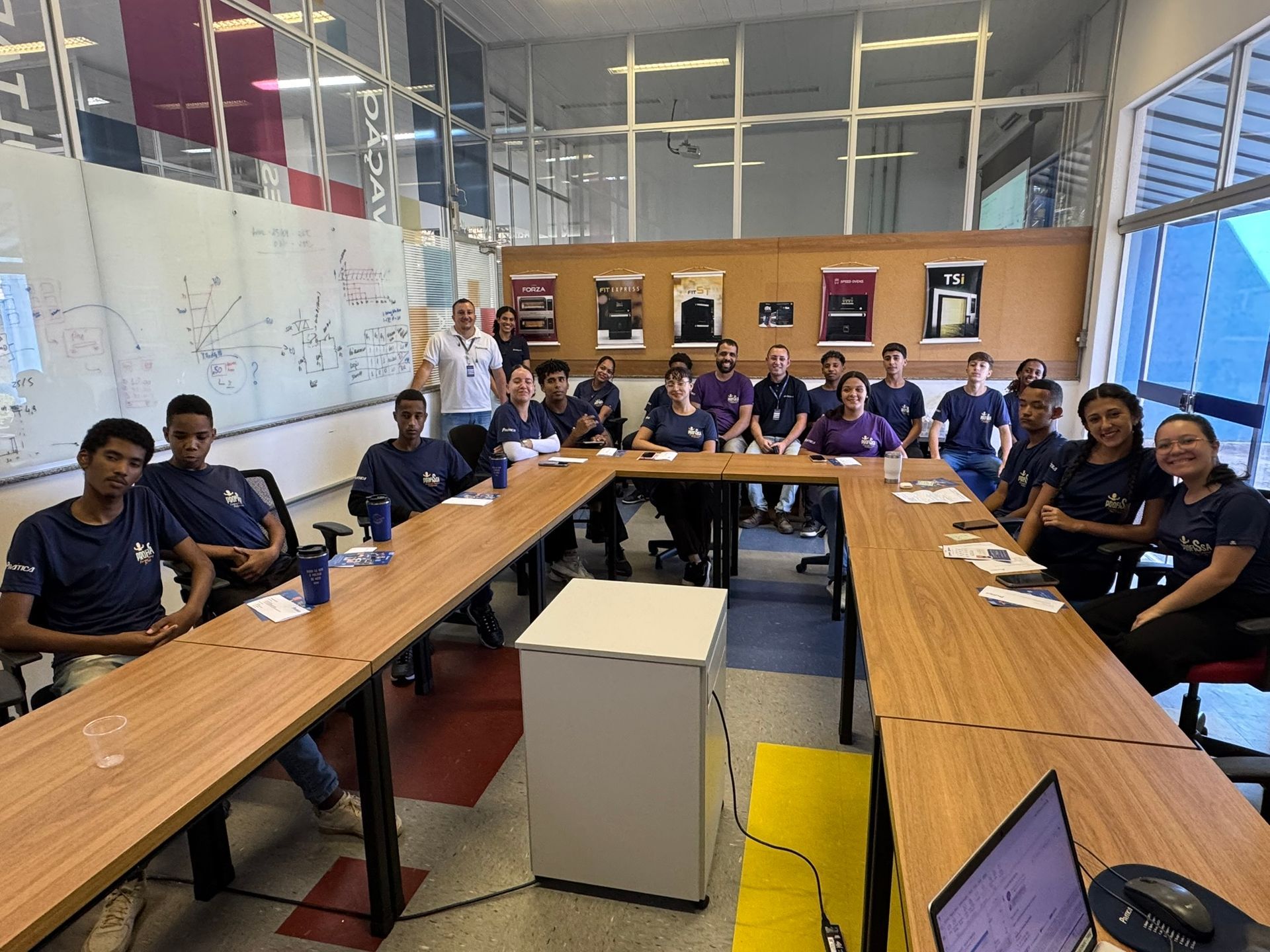 Grupo de pessoas com camisas iguais, sentadas ao redor de uma mesa, dentro de um escritório moderno, olhando para a câmera.