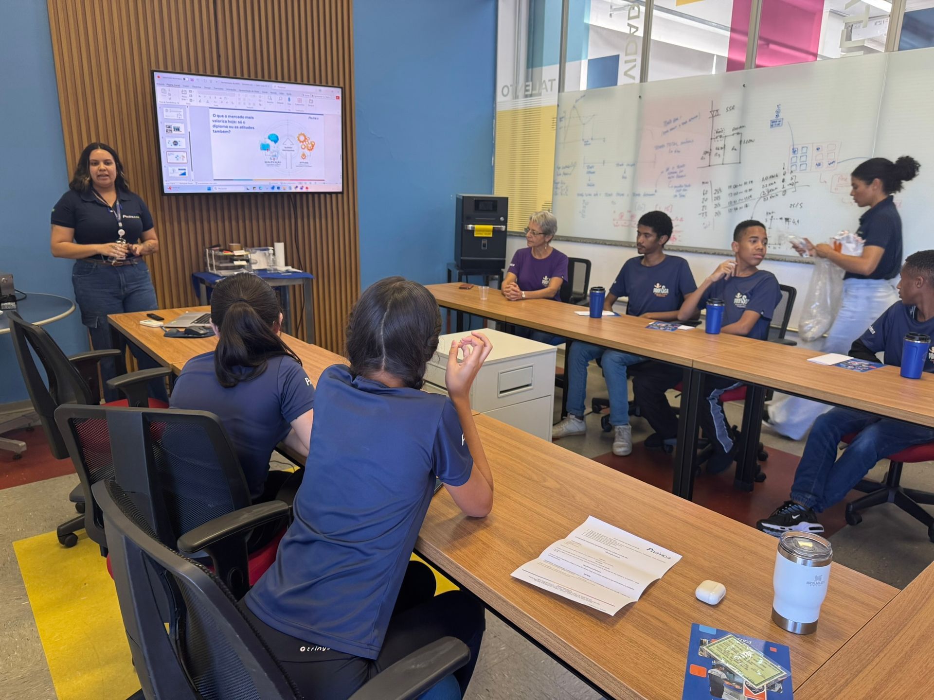 Uma apresentação em sala de aula: pessoas sentadas à mesa assistem a um apresentador falando em frente a uma tela.