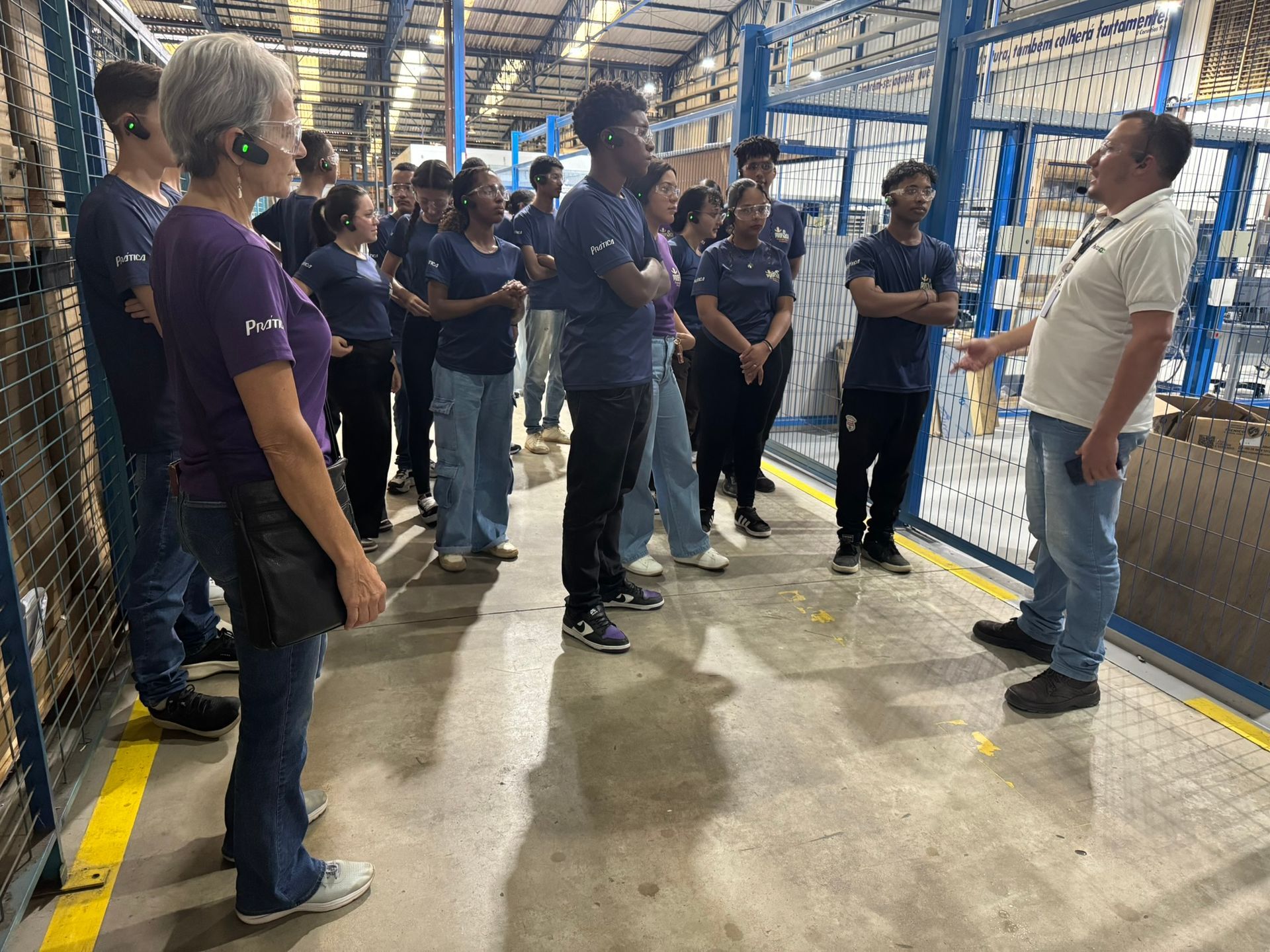 Um grupo de pessoas ouve um homem em um armazém. Eles vestem camisas azuis, e ele aponta para as prateleiras.