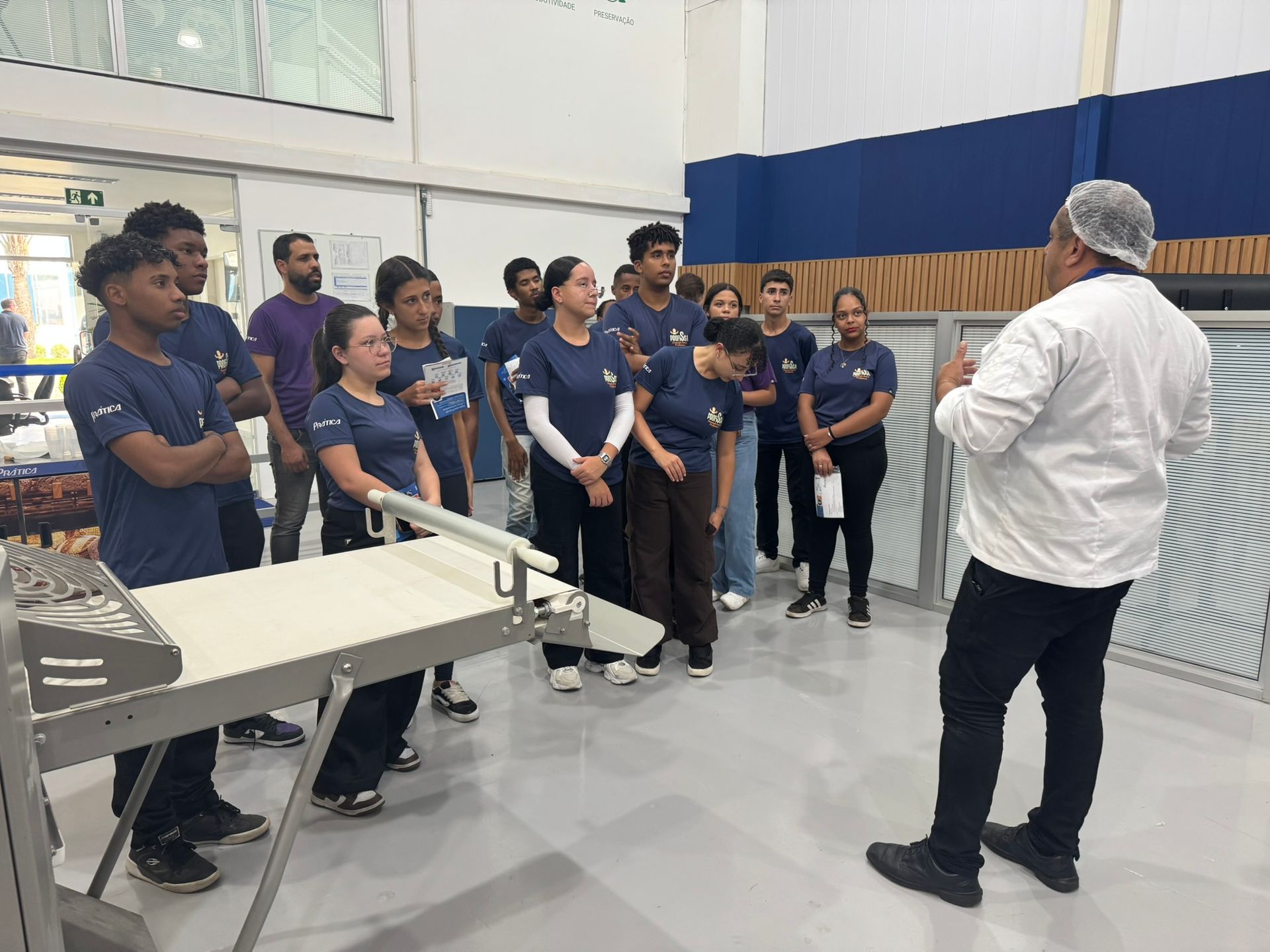 Um grupo de pessoas vestindo camisas azuis ouve um chef em uma área de processamento de alimentos.