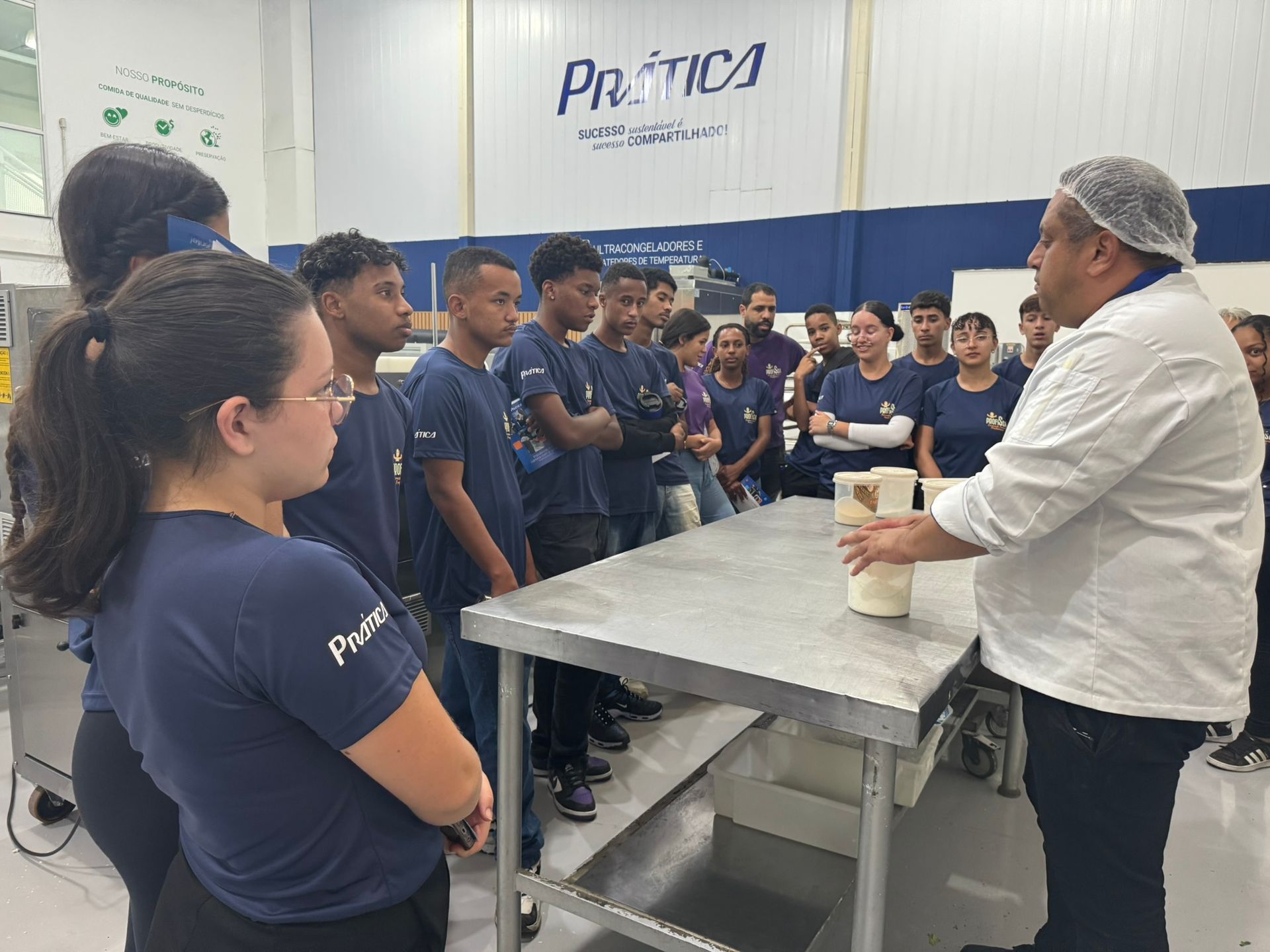 Um chef usando touca de cabelo faz uma demonstração para um grupo de pessoas de camisa azul em uma mesa de aço inoxidável, dentro de uma fábrica.