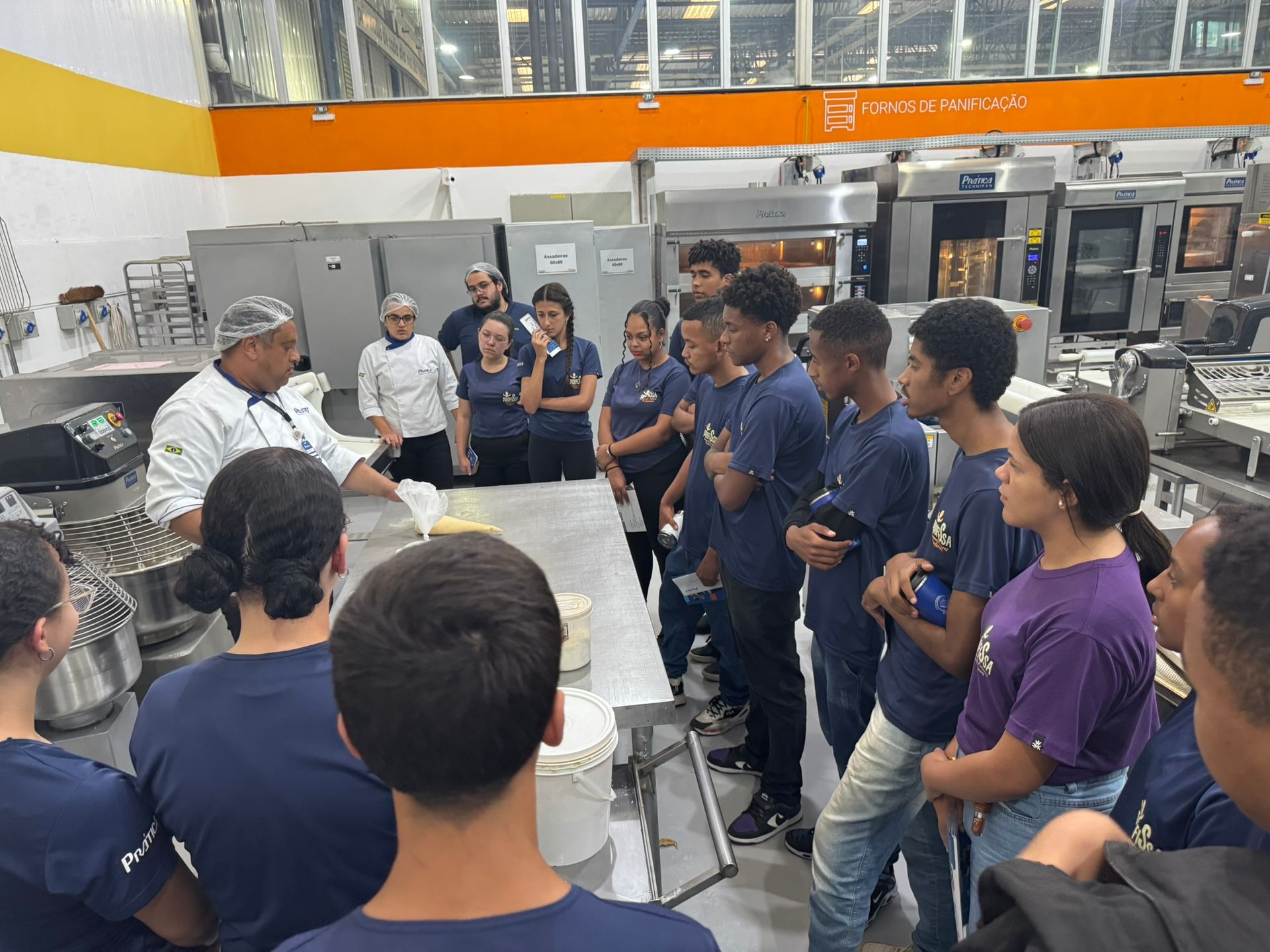 Chef demonstrando técnicas culinárias em uma cozinha profissional para um grupo de alunos vestindo camisas azuis.