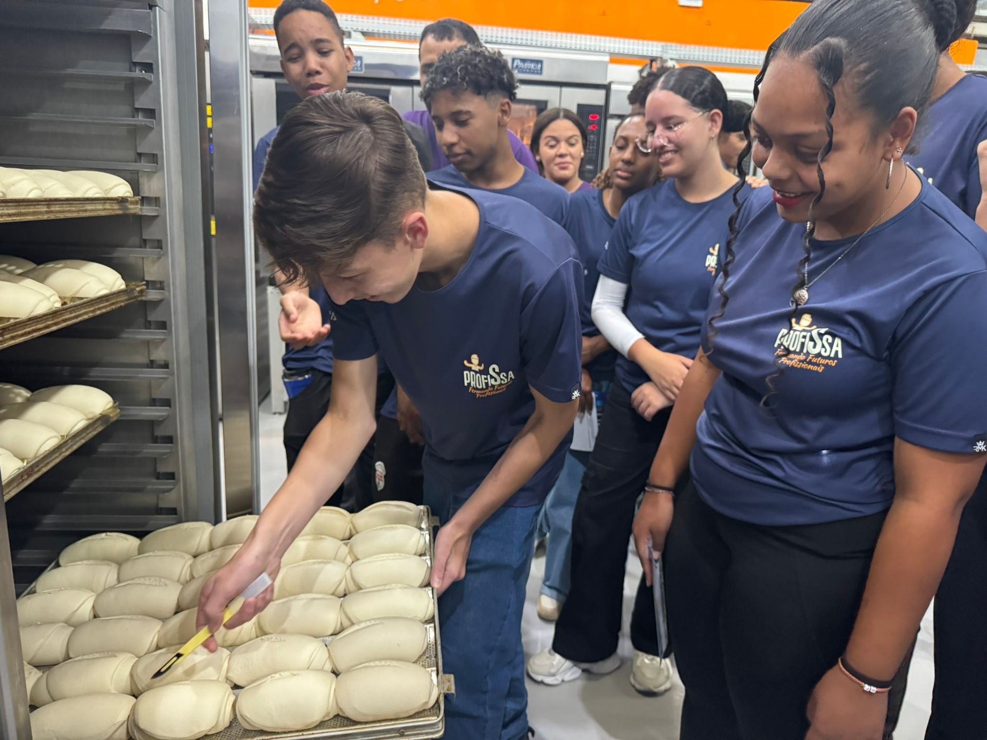 Grupo de estudantes com camisas azuis observando a massa de pão no forno; um dos estudantes a toca com um utensílio.