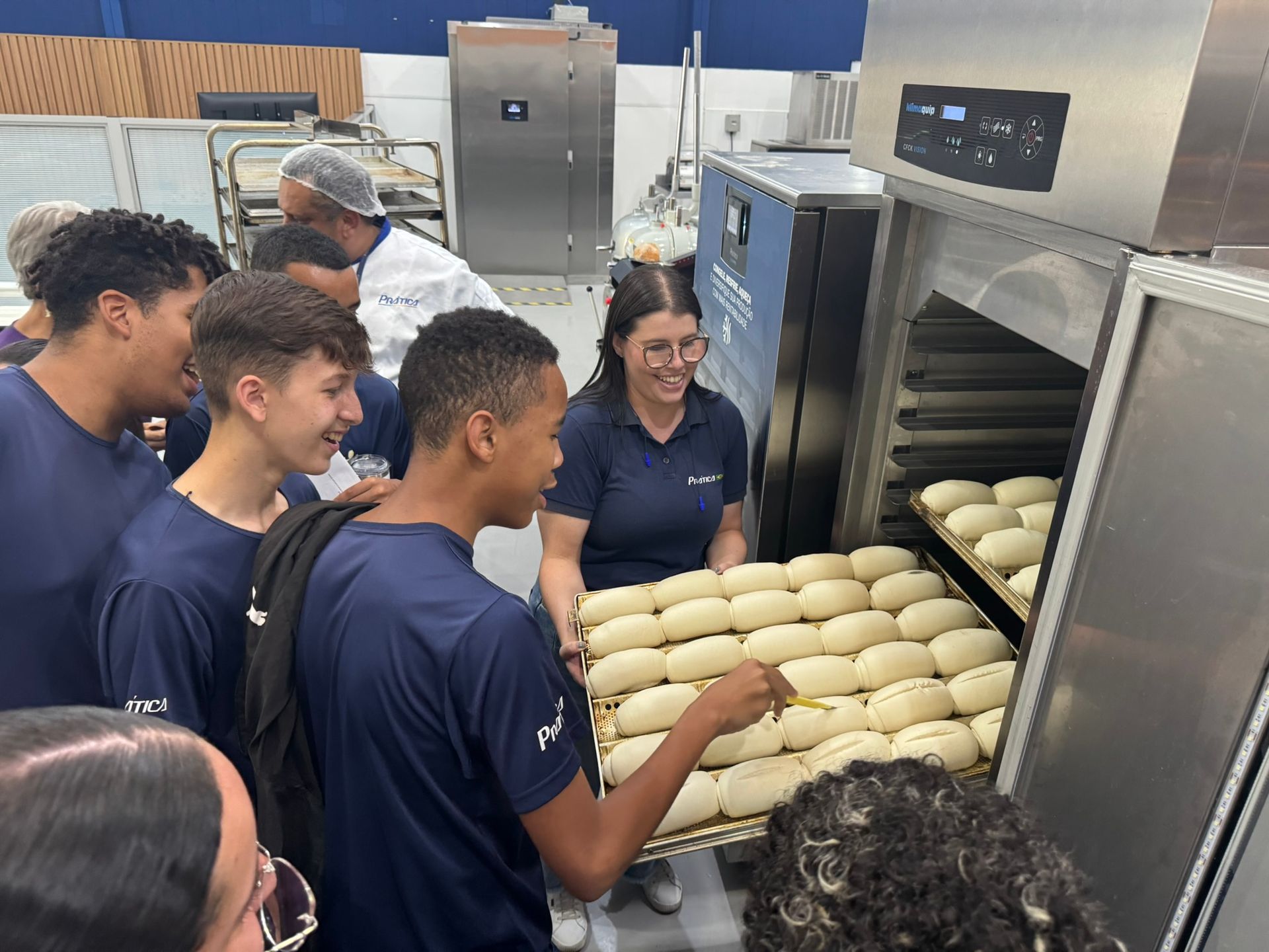 Um grupo observa o processo de assar pão em um forno industrial. Uma mulher segura uma bandeja de pães; outras pessoas sorriem.