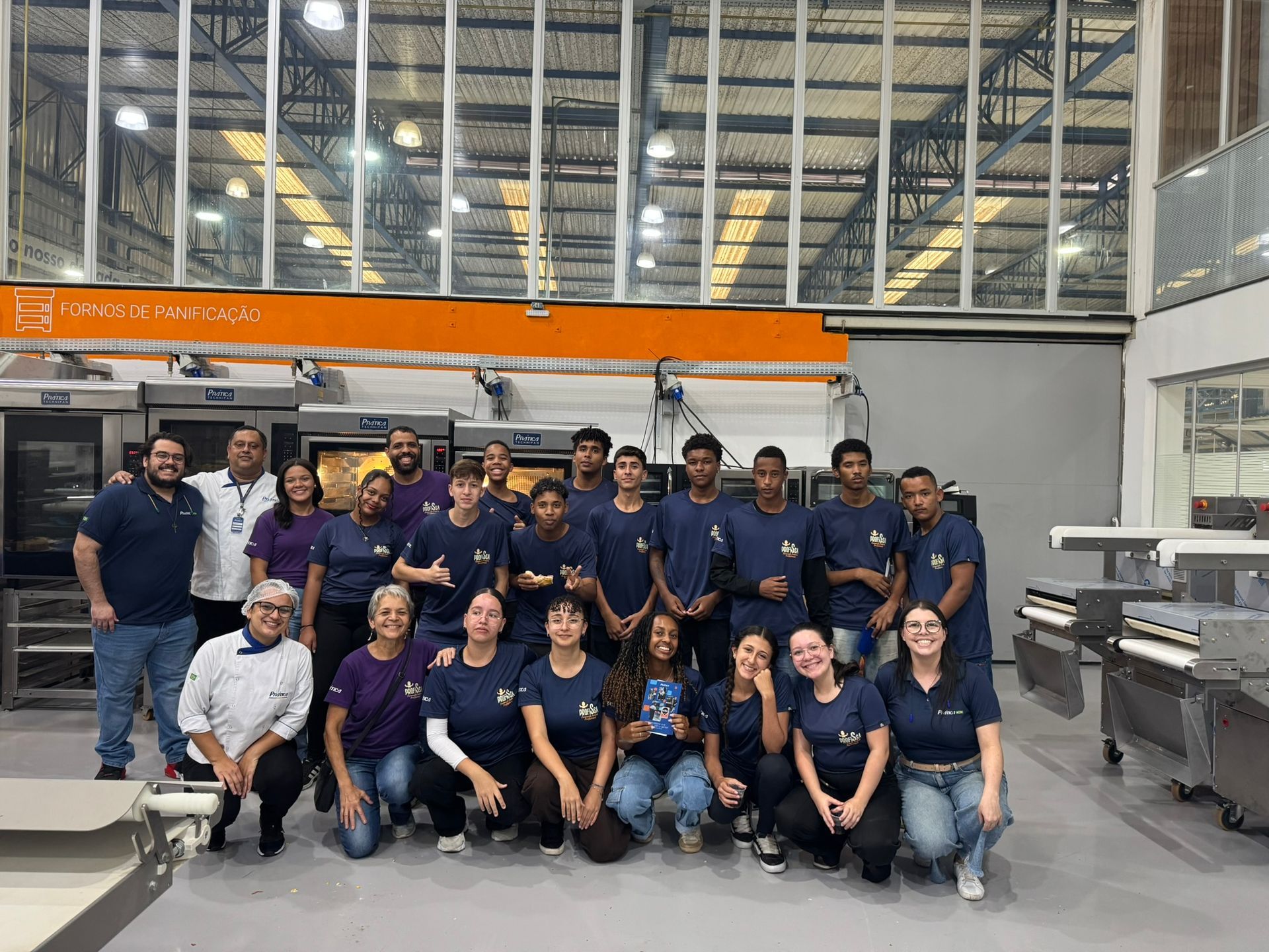 Grupo de pessoas em uma cozinha comercial, posando em frente aos equipamentos; algumas vestindo camisas azuis.