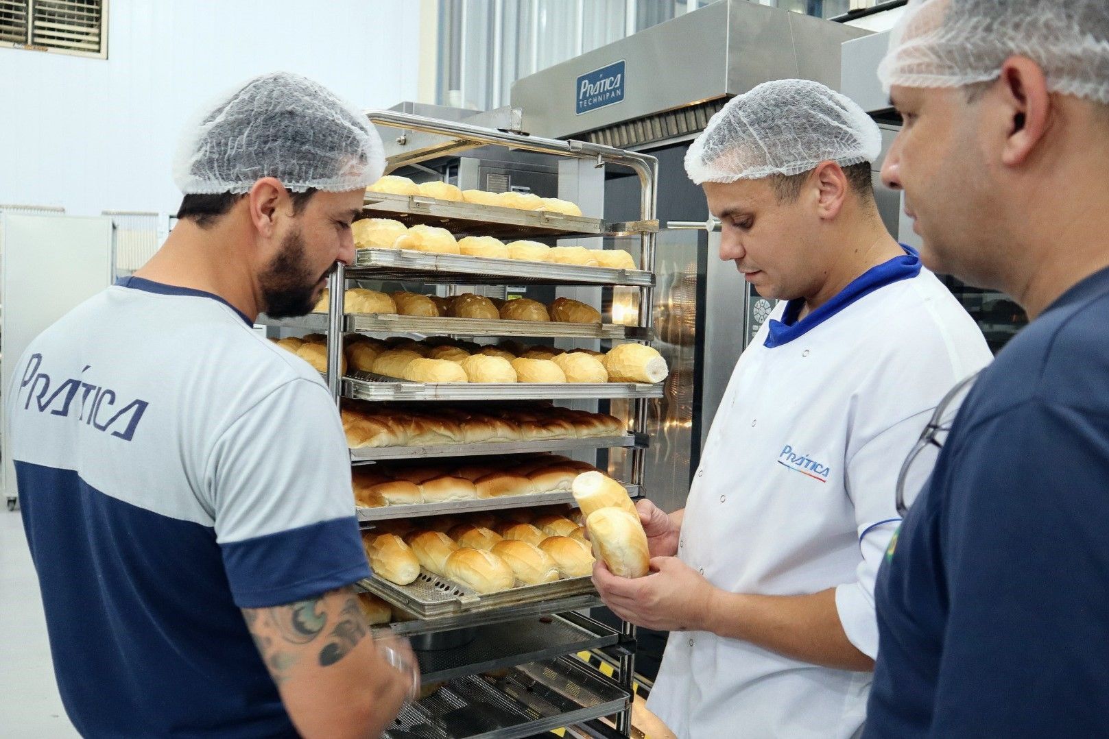Três homens estão trabalhando em uma padaria olhando para pães nas prateleiras.