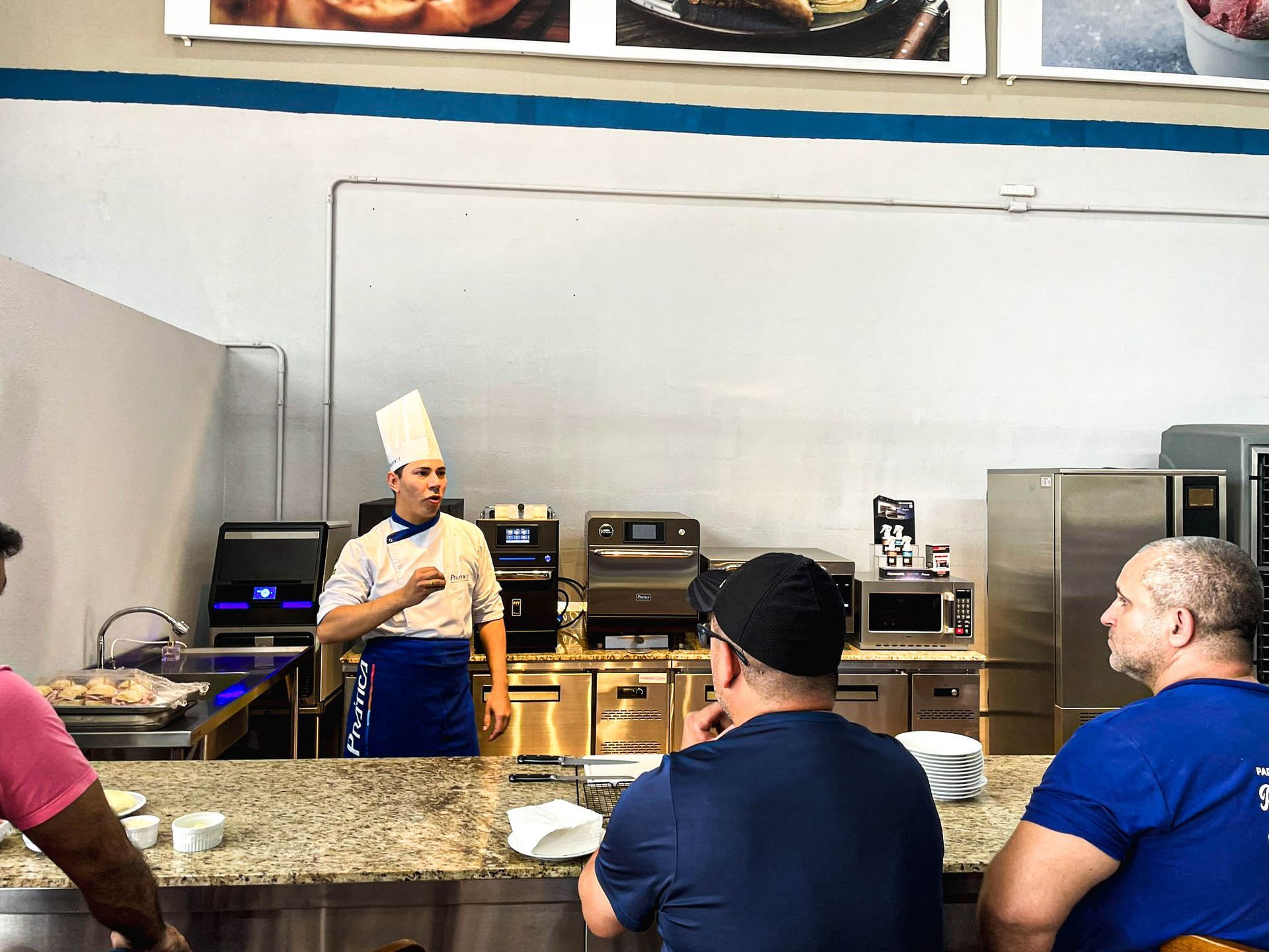 Um homem com chapéu de chef está fazendo uma apresentação para um grupo de pessoas em uma cozinha.