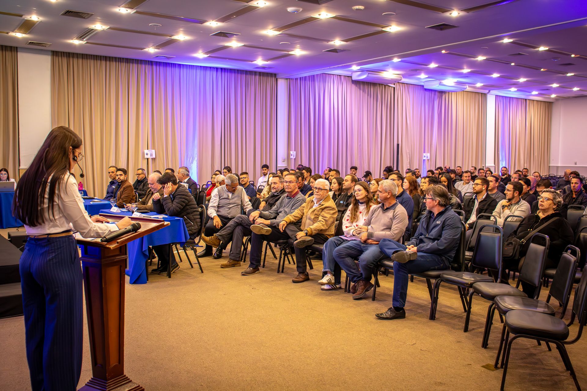 Uma mulher discursa em um pódio para uma plateia em uma sala de conferências. A multidão está sentada, ouvindo.