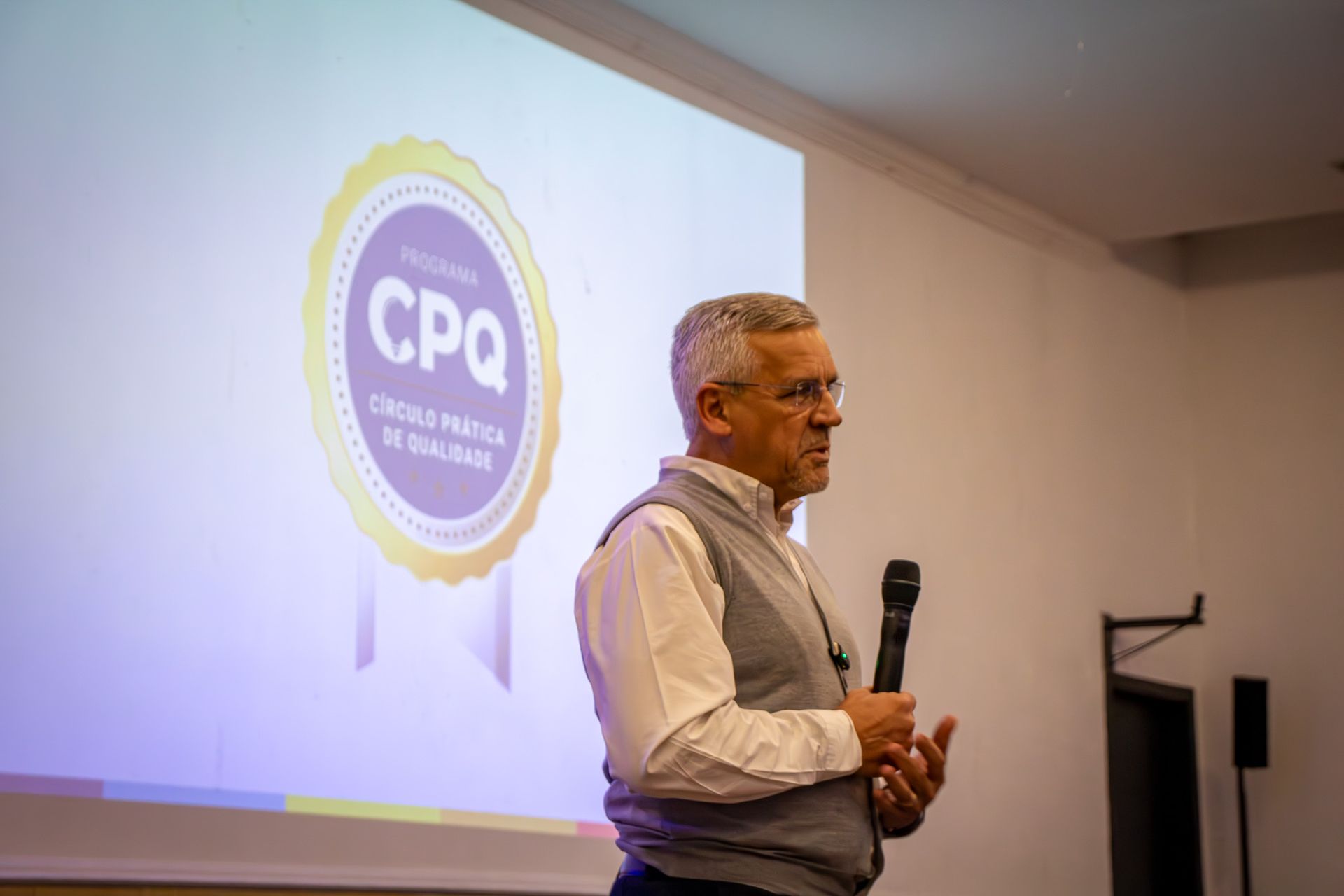 Um homem de cabelos grisalhos fala ao microfone durante uma apresentação. Atrás dele, na tela, há um logotipo com os dizeres