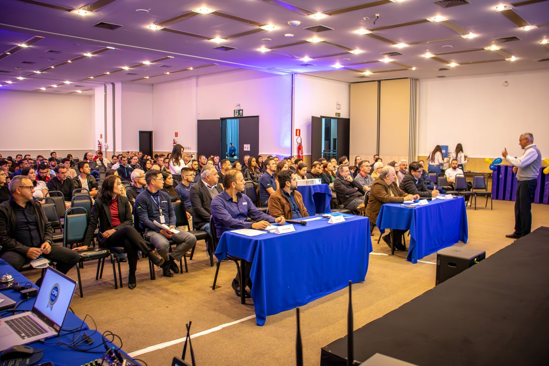 Um homem palestra para um grande público em um salão de conferências. As pessoas sentam-se em mesas e cadeiras, de frente para o palestrante.
