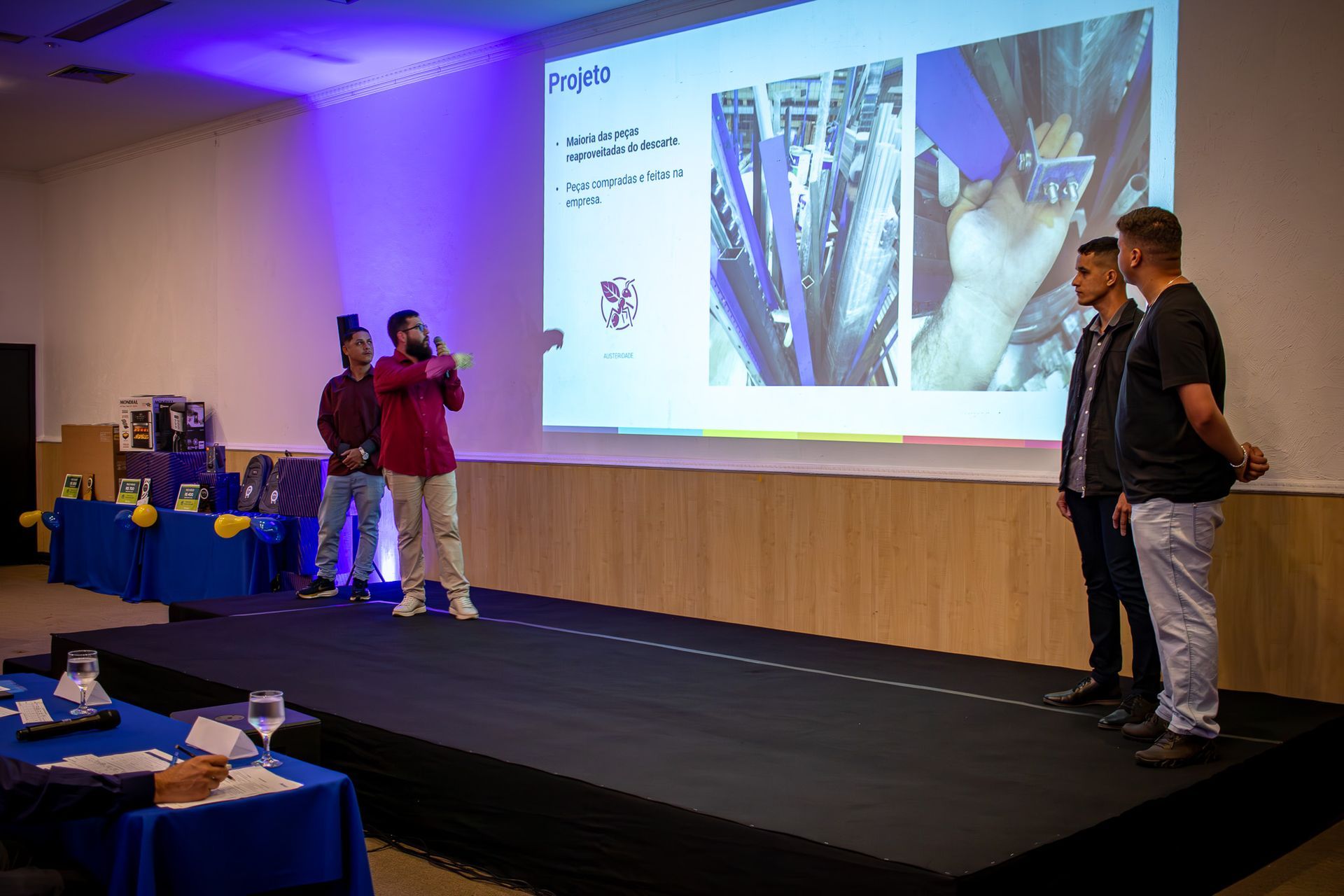 Três homens no palco apresentando-se em uma conferência, um deles falando ao microfone. Uma tela de projetor exibe gráficos.
