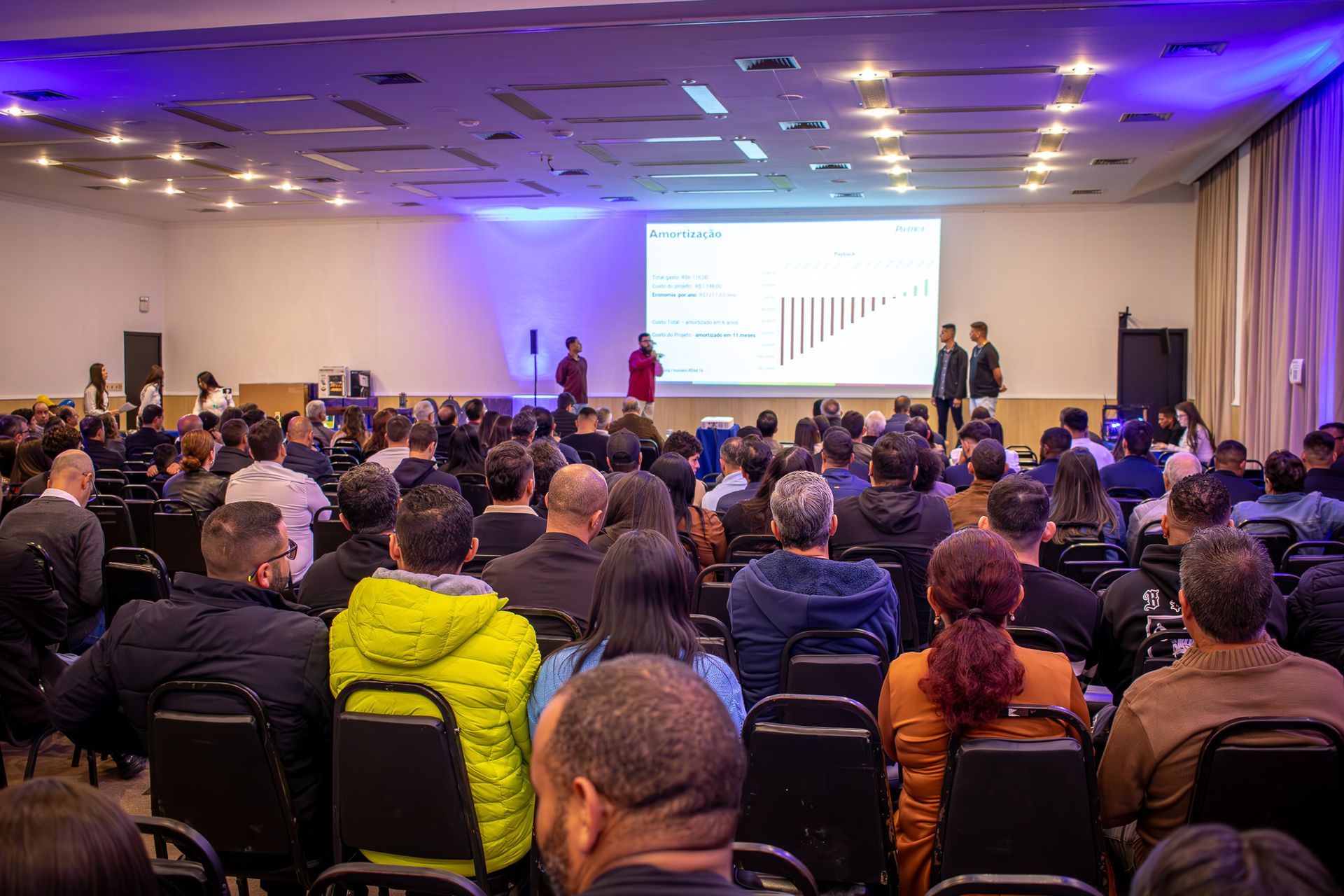 Uma grande sala de conferências com uma plateia diante de uma apresentação em uma tela, dois apresentadores no palco e participantes sentados.