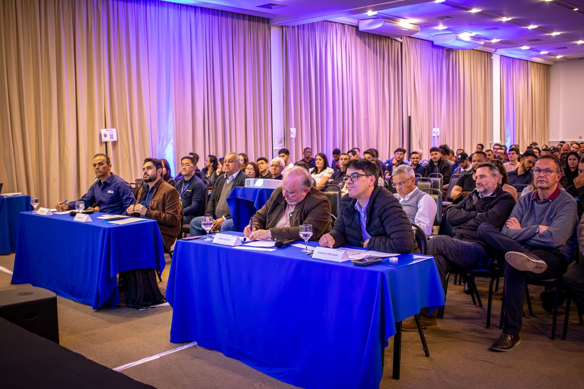 Público em uma conferência com palestrantes em mesas. Toalhas de mesa azuis, cortinas bege e um esquema de cores neutro.