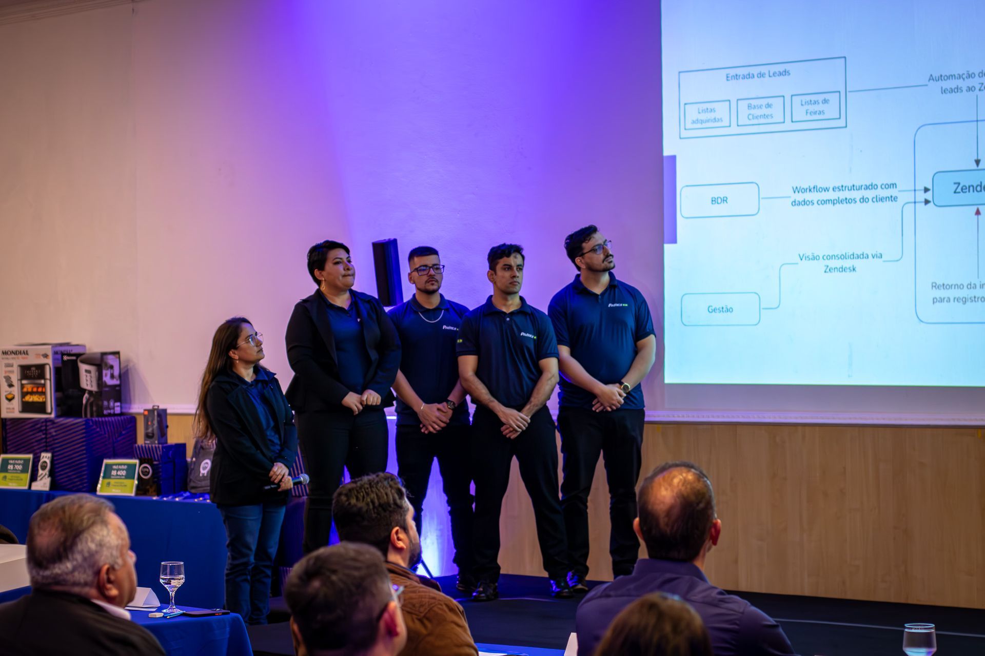 Uma apresentação em um ambiente de conferência. Um grupo de cinco pessoas de camisa azul está no palco, observando um diagrama exibido em uma tela. O público assiste.