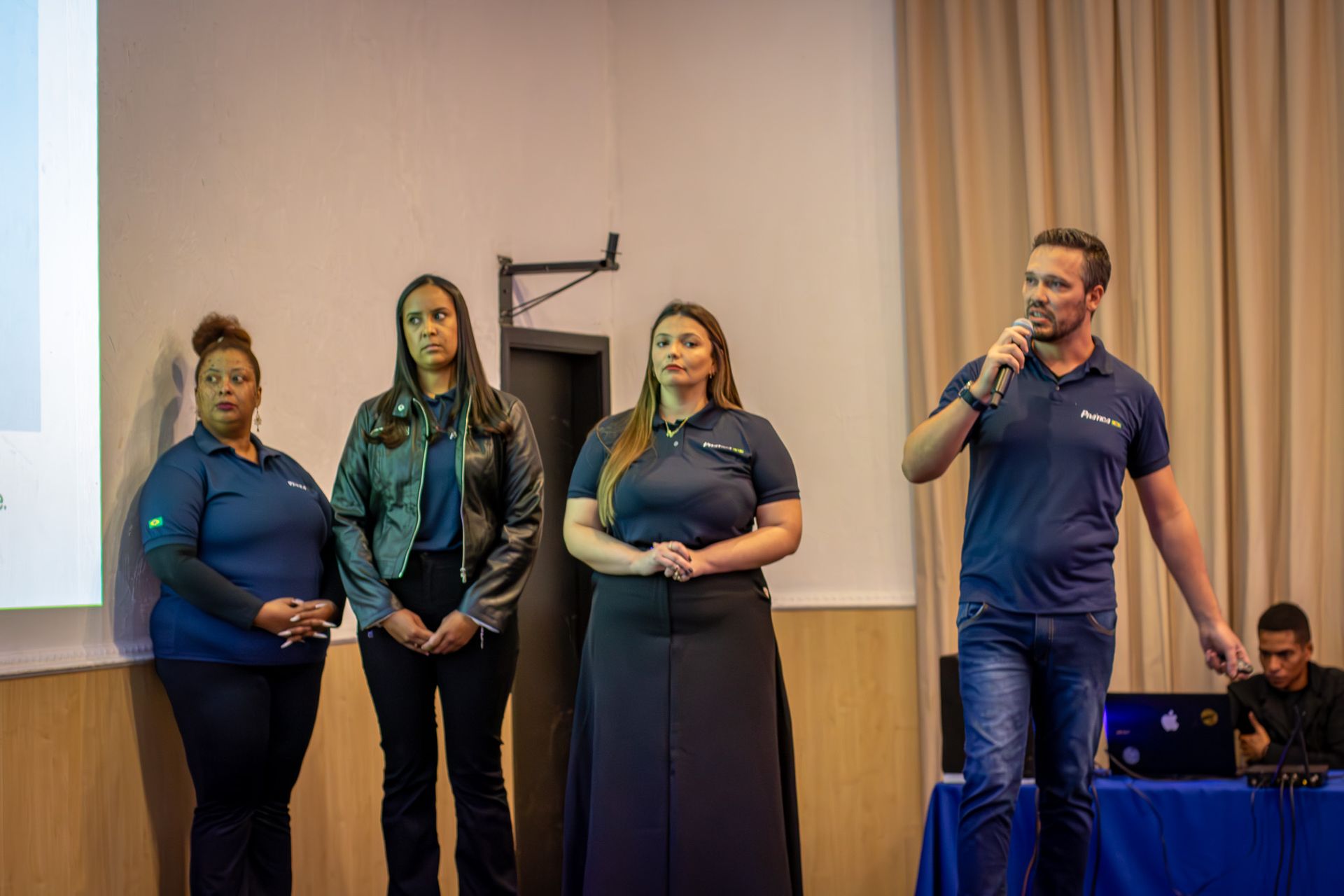 Quatro pessoas no palco durante uma apresentação. Um homem com um microfone fala; três mulheres estão de pé ao lado, todas vestindo camisas azul-marinho.