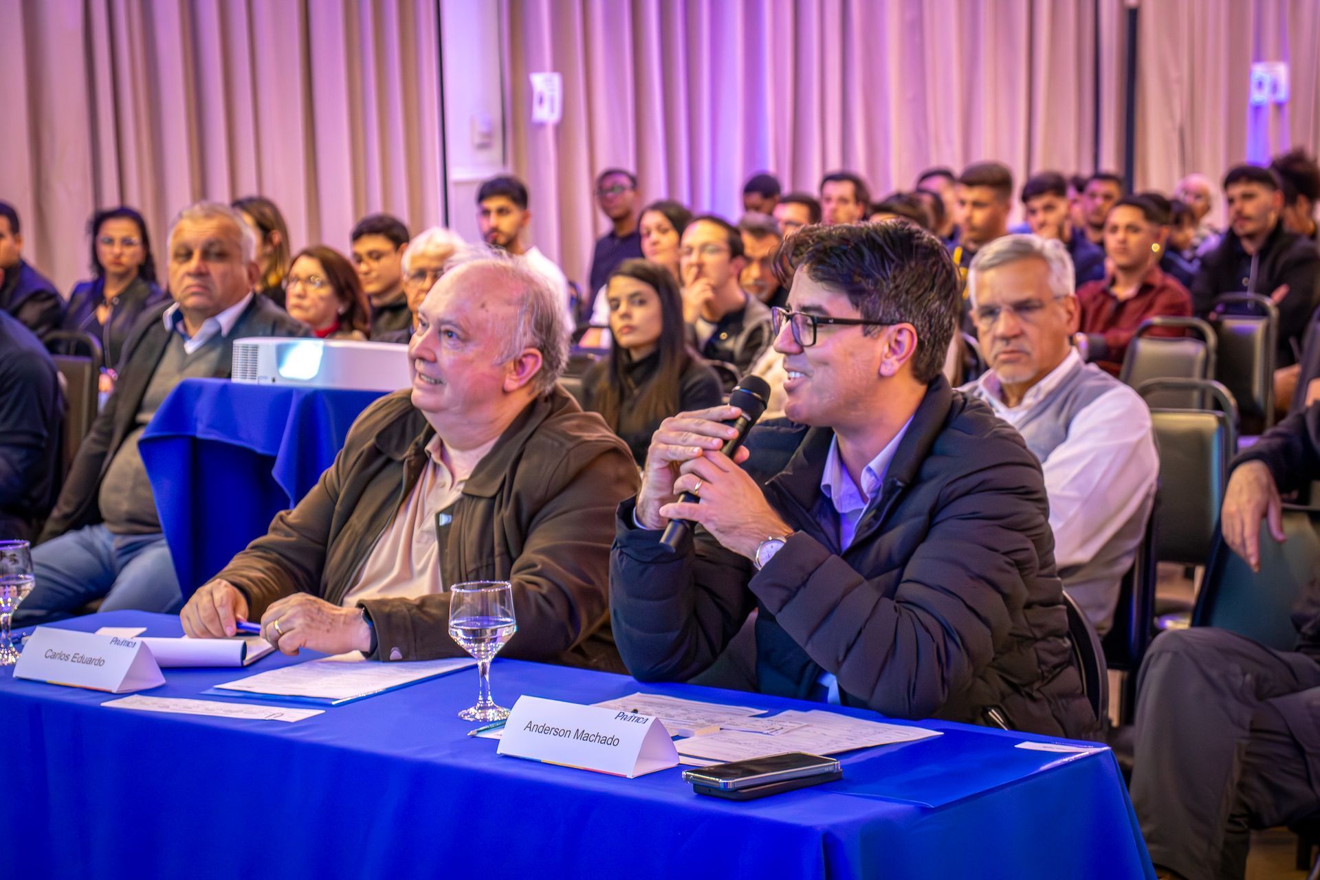 Homens discursando em uma mesa de conferência diante de uma plateia. A sala está iluminada com luz roxa.