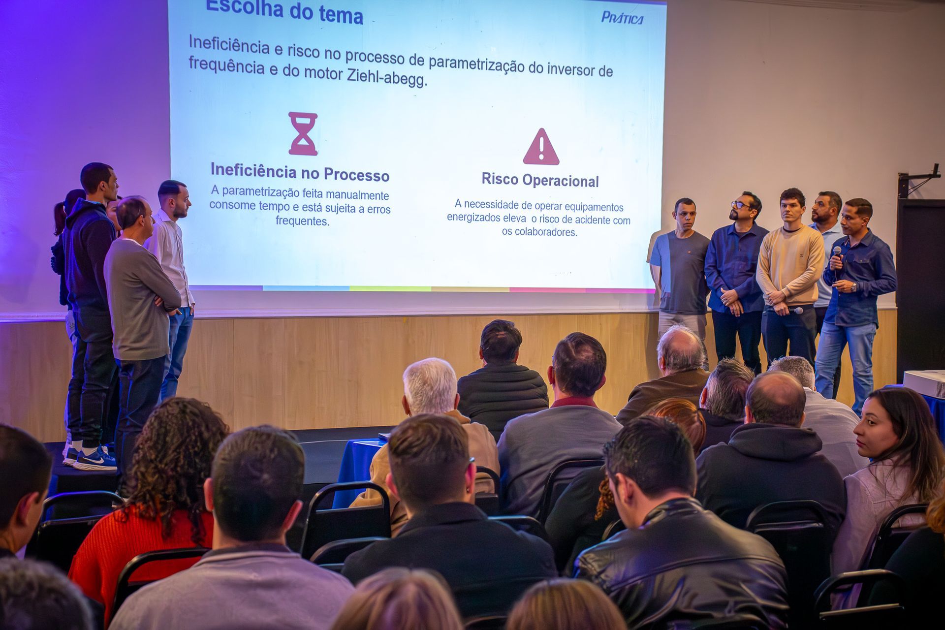 Uma apresentação em uma sala de conferências. Os palestrantes ficam diante de um slide projetado enquanto o público assiste. O slide contém texto e gráficos.