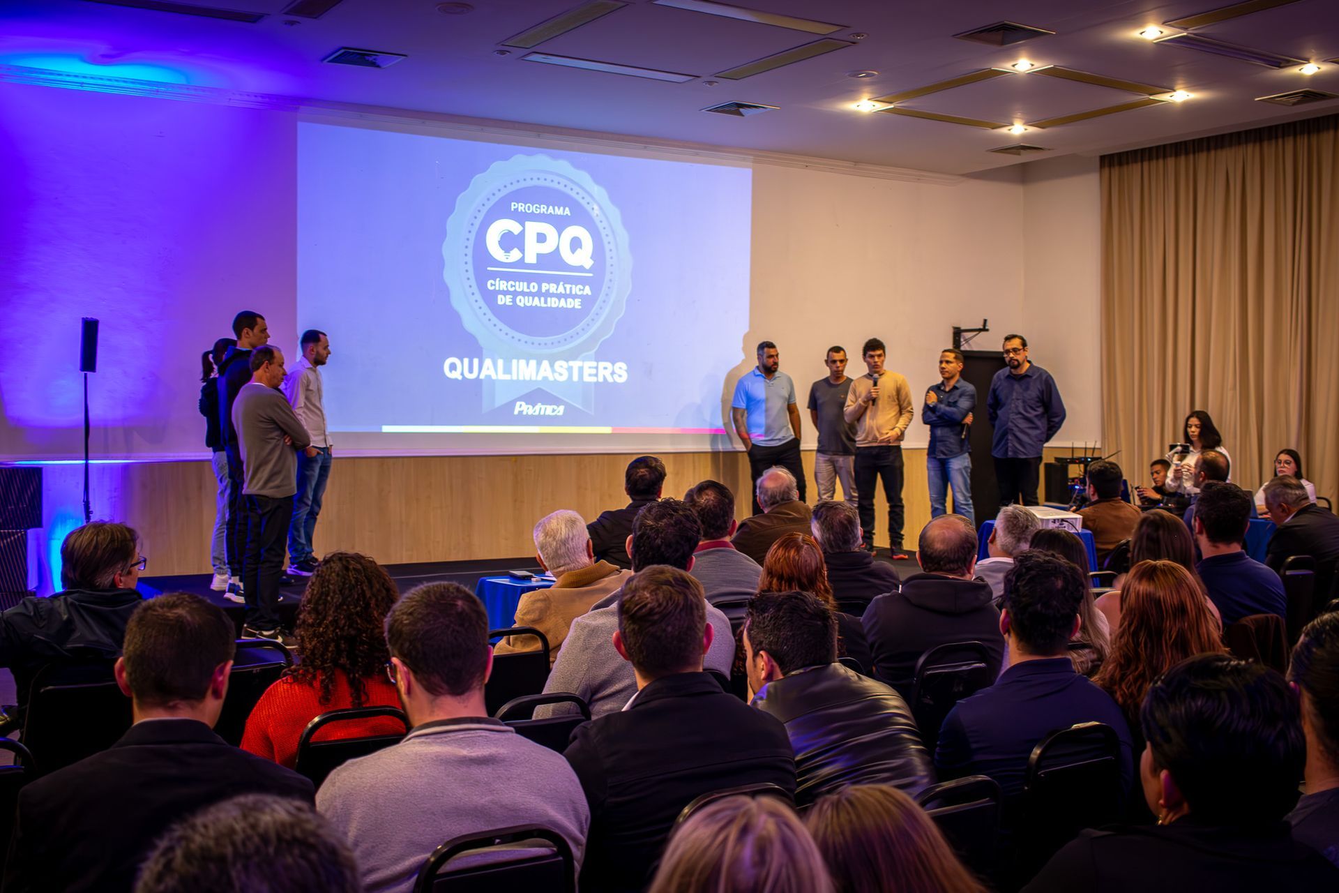 Uma apresentação em uma sala de conferências com um grupo de homens em um palco, falando para uma plateia sentada. Um projetor exibe texto.