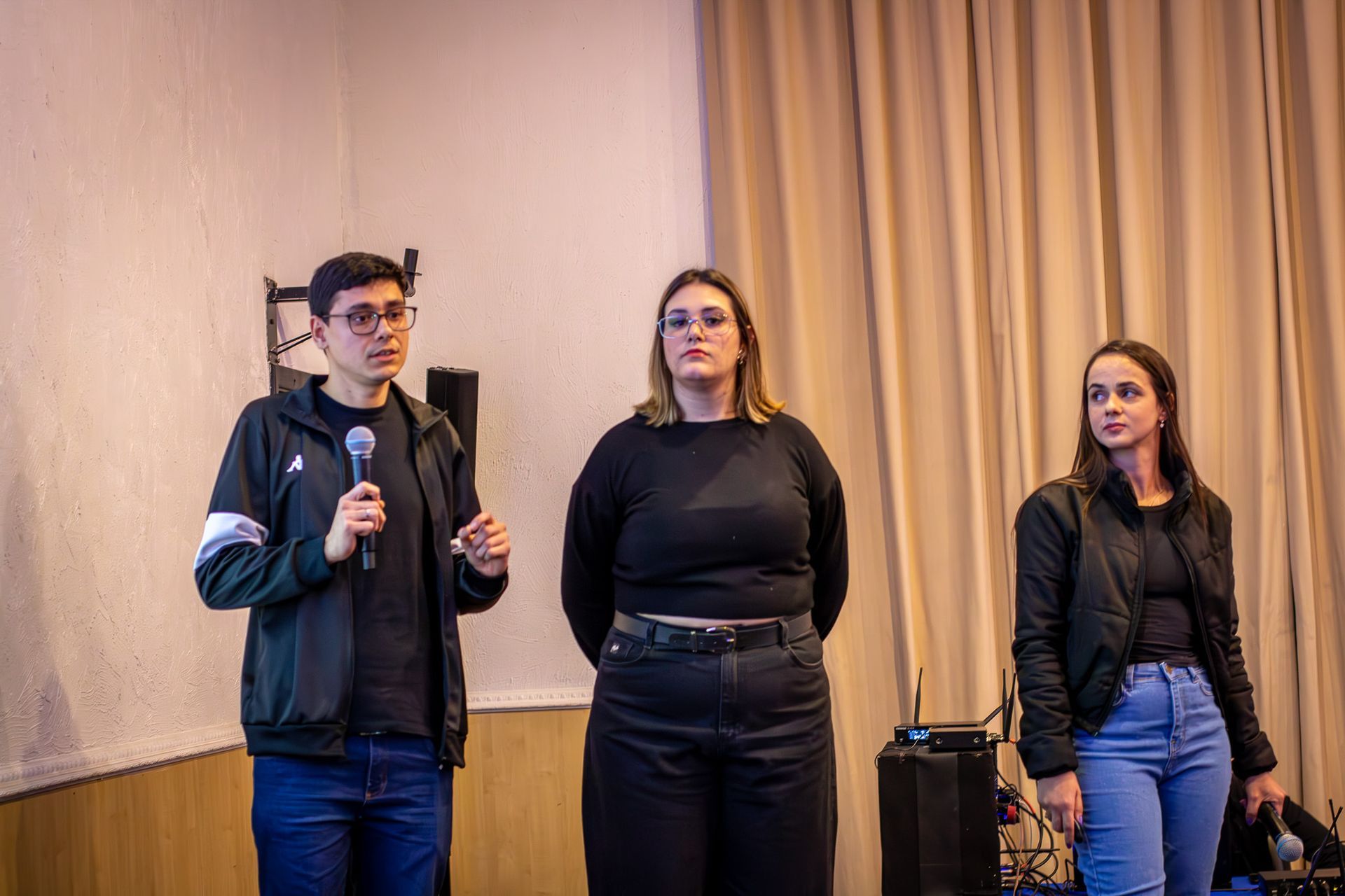 Três pessoas em um palco, duas mulheres e um homem, falando em microfones. Eles parecem estar fazendo uma apresentação em uma sala com cortinas bege.