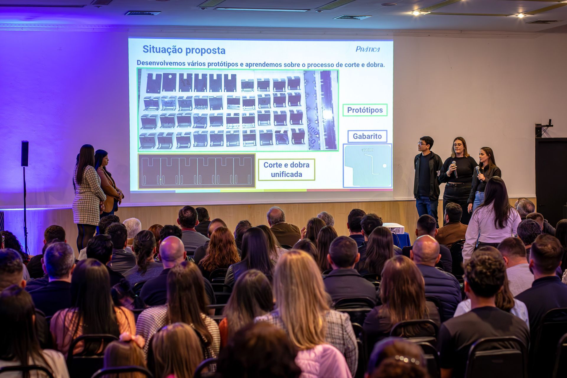 Uma apresentação em um ambiente de conferência com palestrantes em um palco, um slide projetado e um grande público.