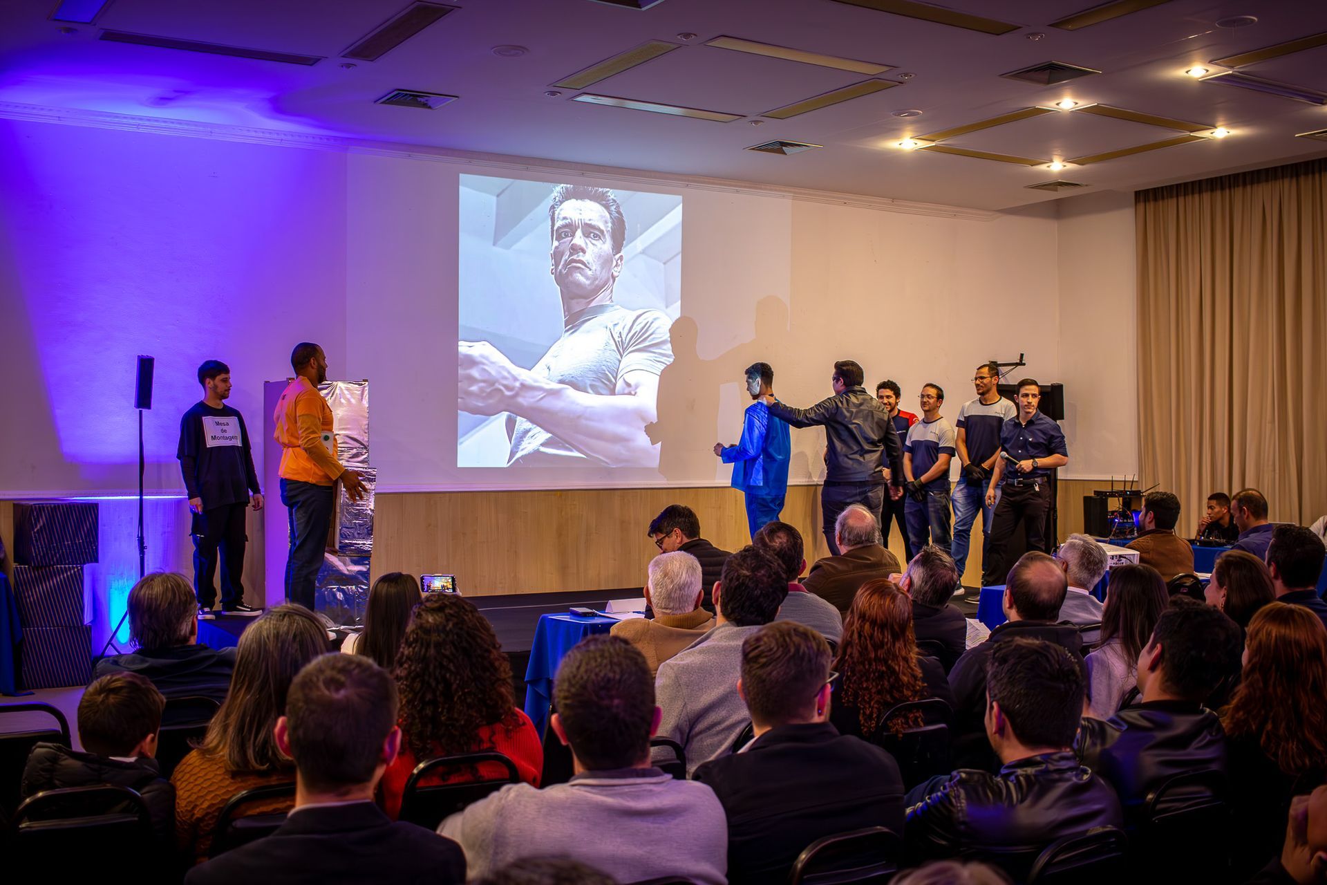 Um grupo de pessoas no palco durante uma apresentação, com um projetor exibindo uma foto. Uma plateia sentada à frente.
